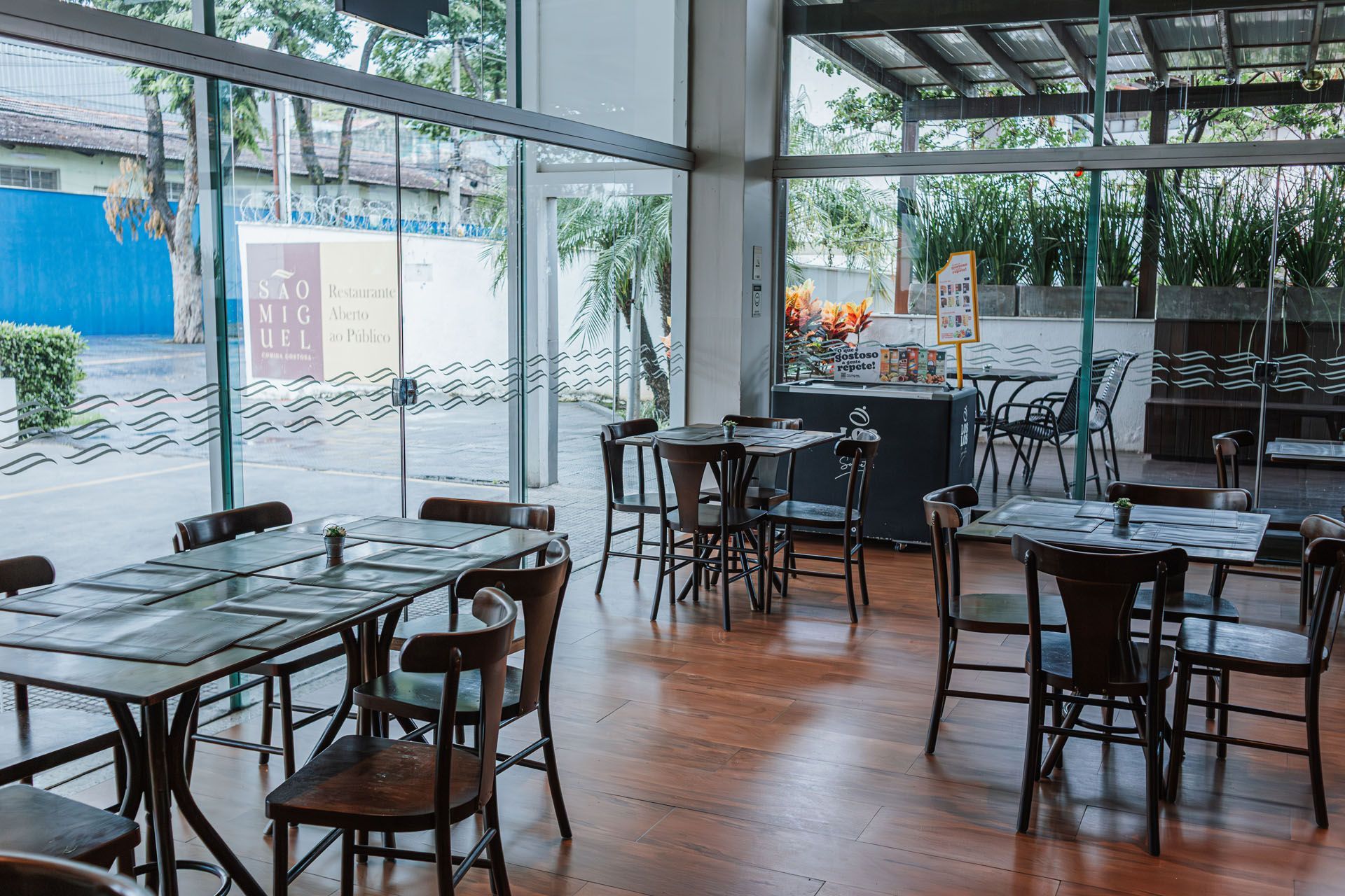 Café vazio com mesas e cadeiras de madeira junto a grandes janelas de vidro, com vista para um dia chuvoso.