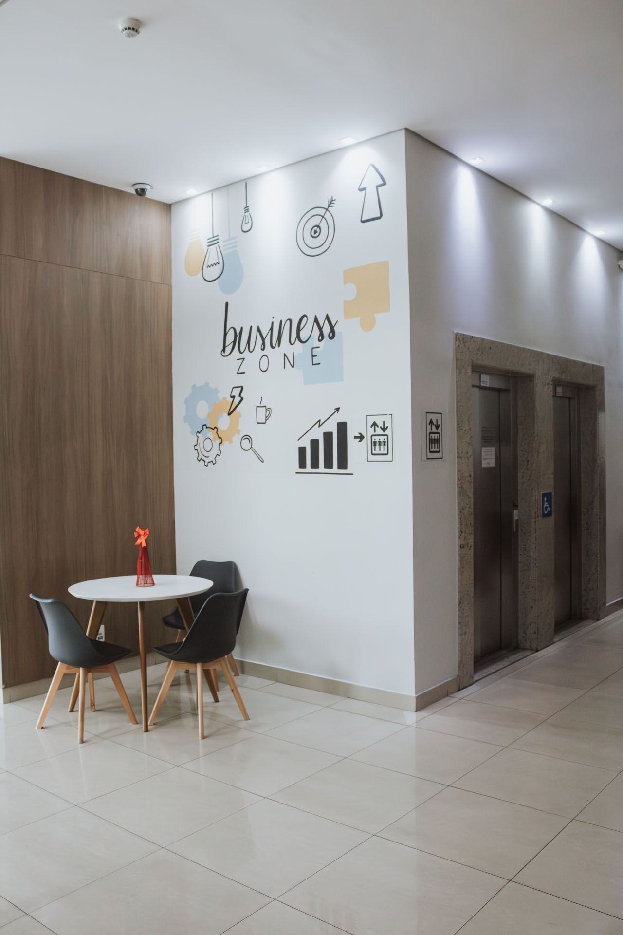 Hall de entrada moderno com pequena mesa redonda, cadeiras, obras de arte na parede e portas de elevador.