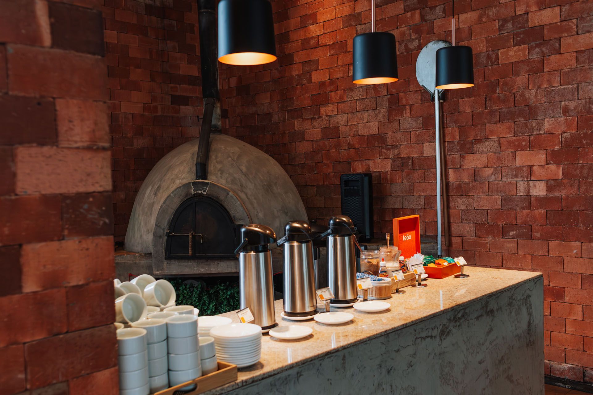 Balcão de buffet com cafeteiras, xícaras, doces e frutas ao lado de um forno de tijolos em um ambiente de restaurante aconchegante.