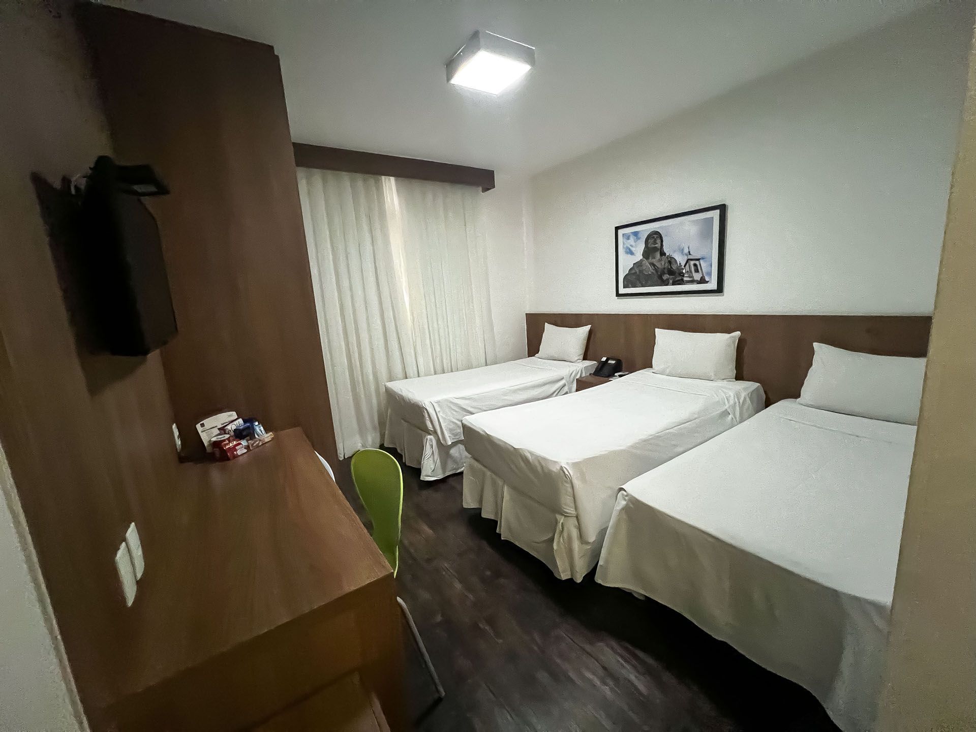 Quarto de hotel com três camas de solteiro arrumadas, escrivaninha, TV e janela com cortinas.