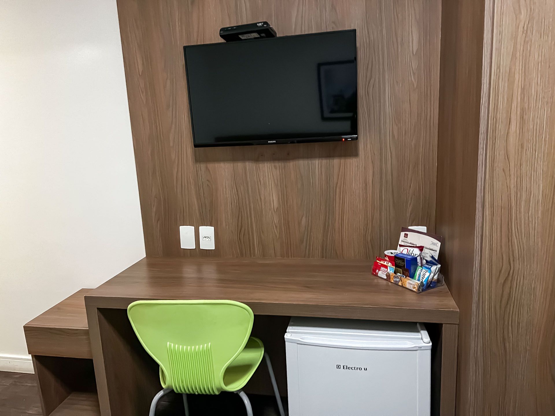 Pequena escrivaninha de hotel com cadeira verde, TV fixada na parede, frigobar e lanches em um painel de madeira.