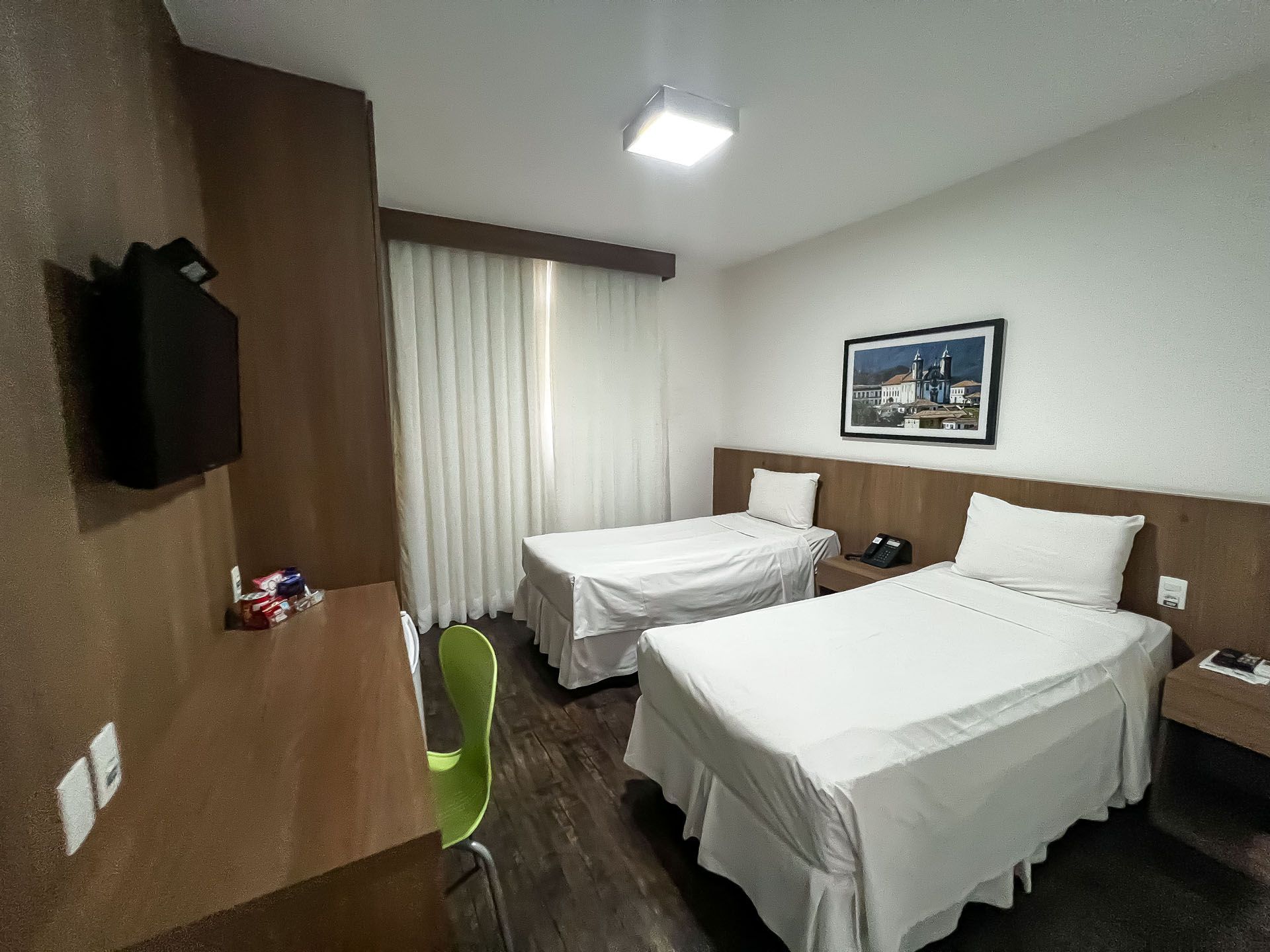Quarto de hotel com duas camas de solteiro, escrivaninha, cadeira verde e cortinas na janela.