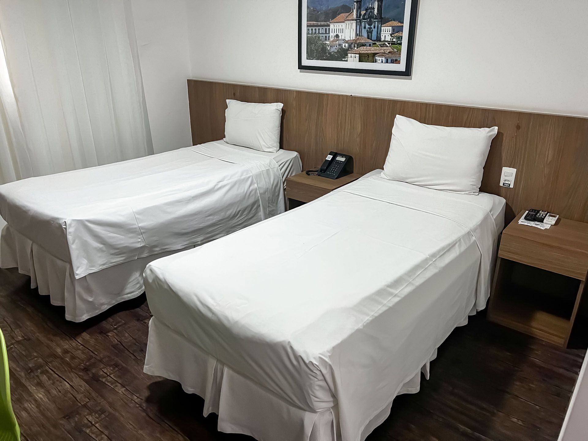 Quarto de hotel com duas camas de solteiro, lençóis brancos, cabeceira de madeira e mesas de cabeceira.