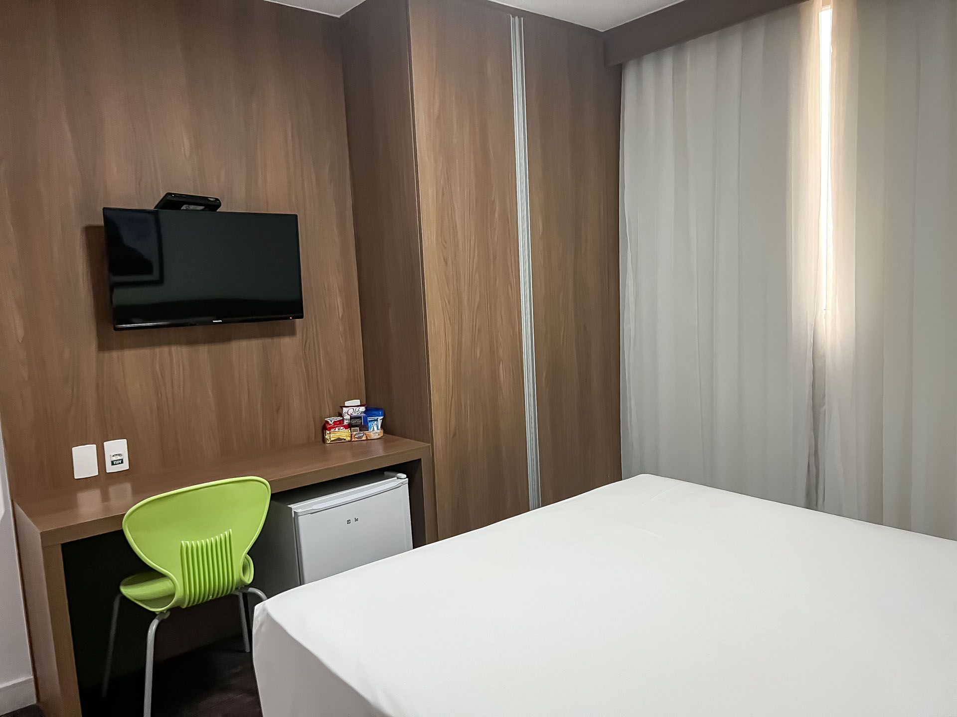 Quarto de hotel pequeno com cama, escrivaninha, cadeira verde, TV de parede e janela com cortina.