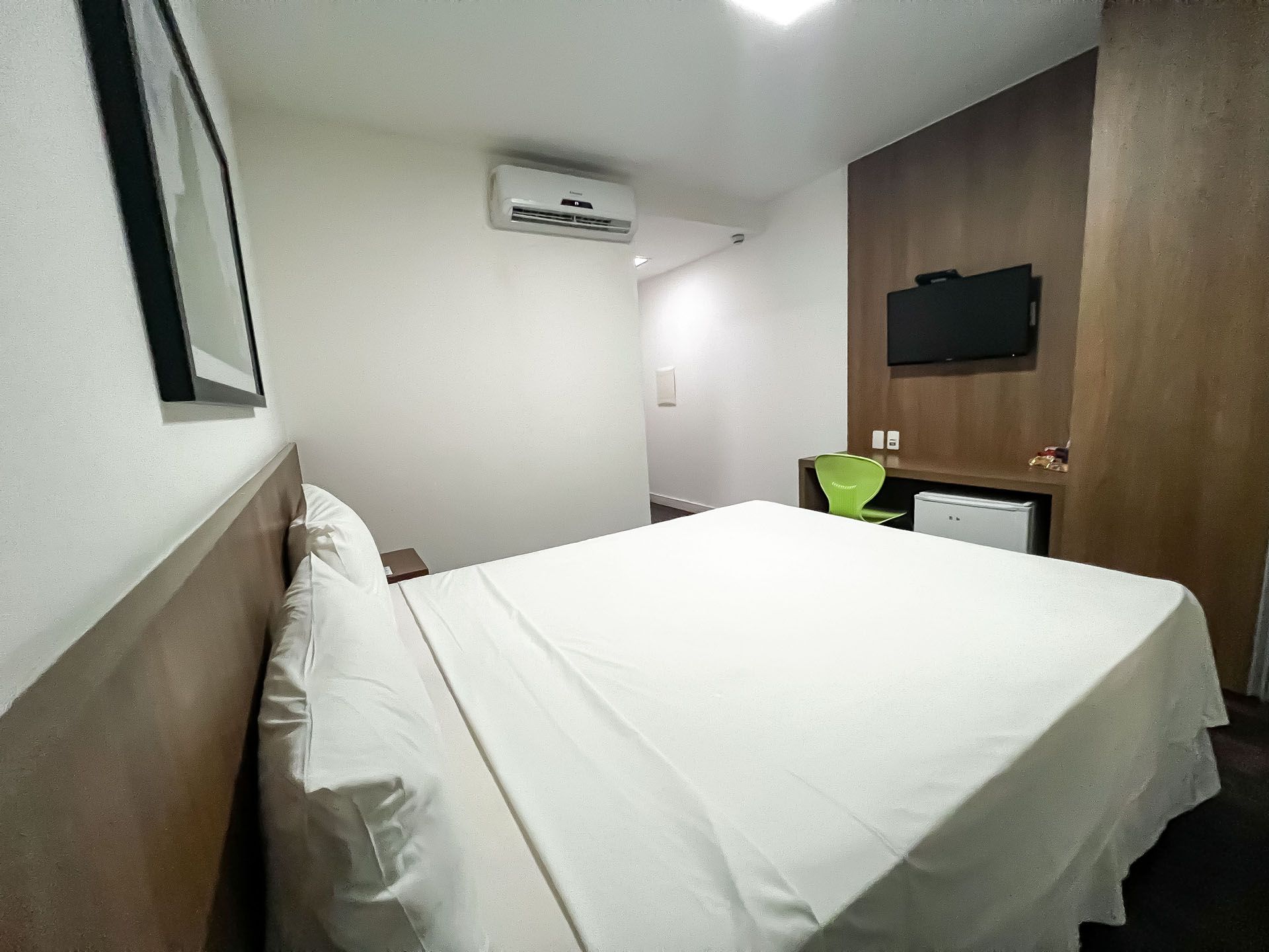 Quarto de hotel pequeno com uma cama branca desarrumada, TV de parede, ar-condicionado e parede com painéis de madeira como destaque.