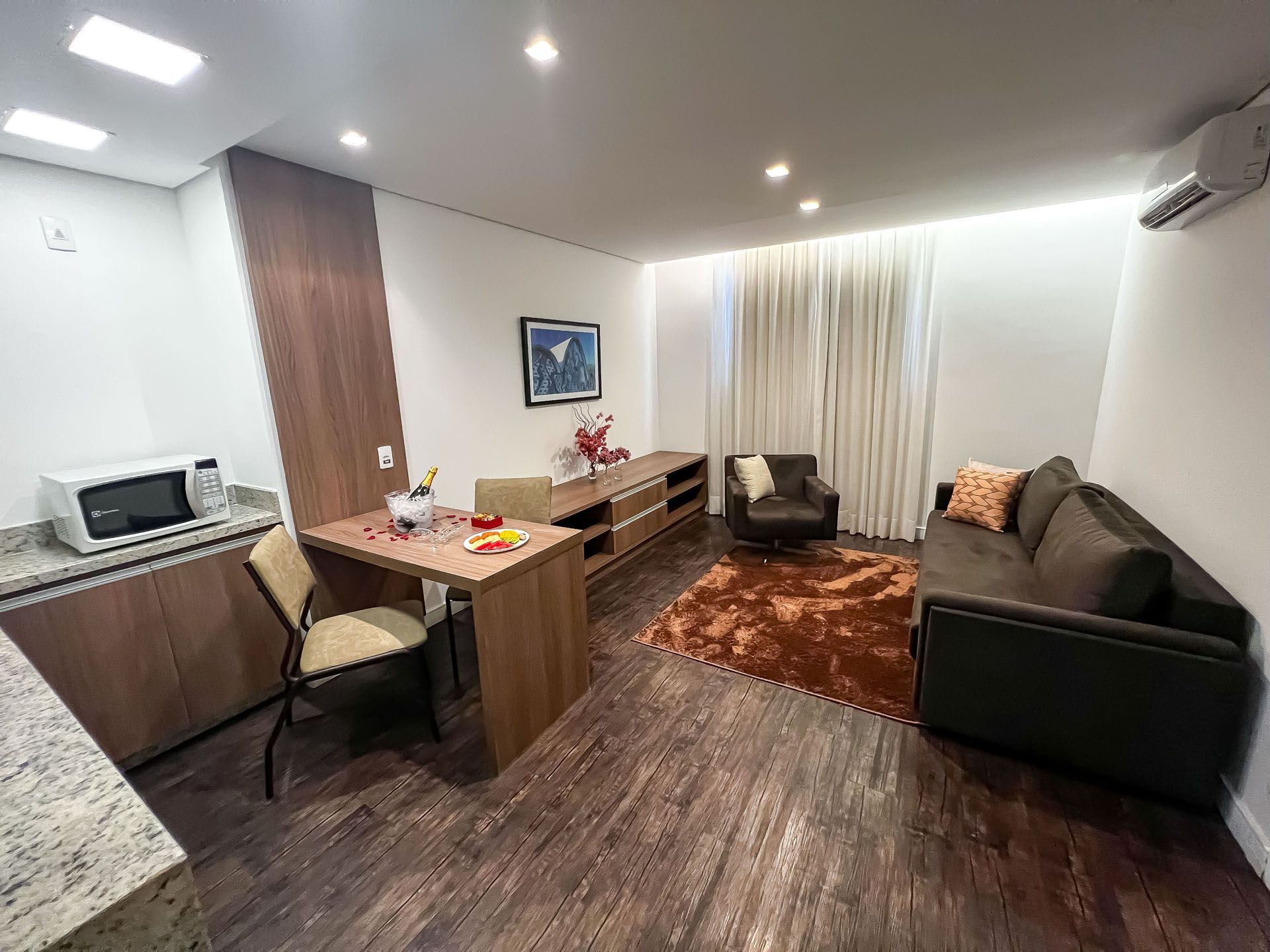 Sala de estar compacta com sofá, cadeiras, escrivaninha pequena, piso de madeira e janela com cortina.