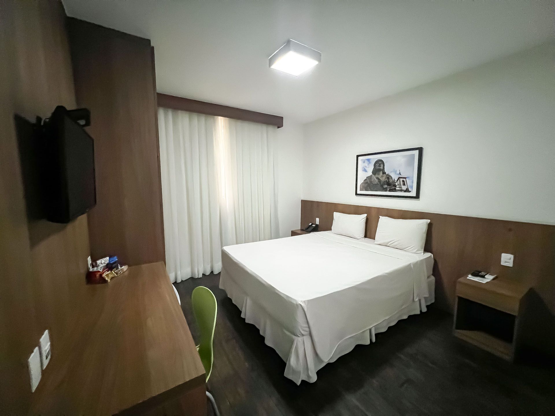 Quarto de hotel pequeno com cama, escrivaninha, cadeira, TV de parede e janela com cortinas.
