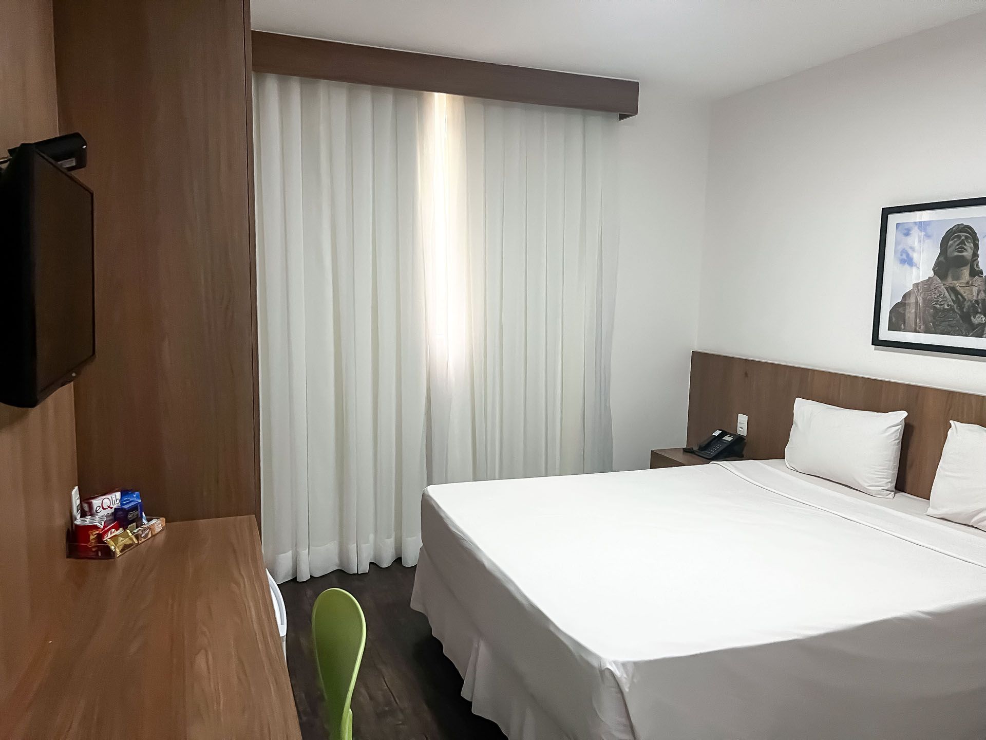 Quarto de hotel moderno com cama bem arrumada, cortinas, TV de parede e área de trabalho.