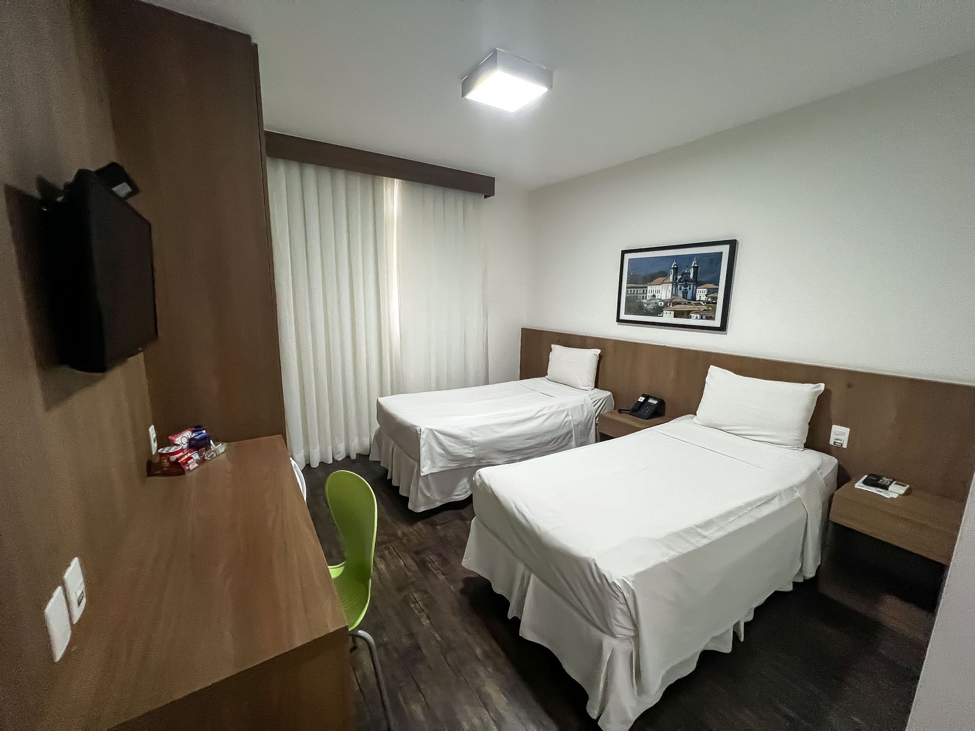 Quarto de hotel pequeno com duas camas de solteiro, paredes revestidas de madeira, uma escrivaninha e uma janela com cortinas.