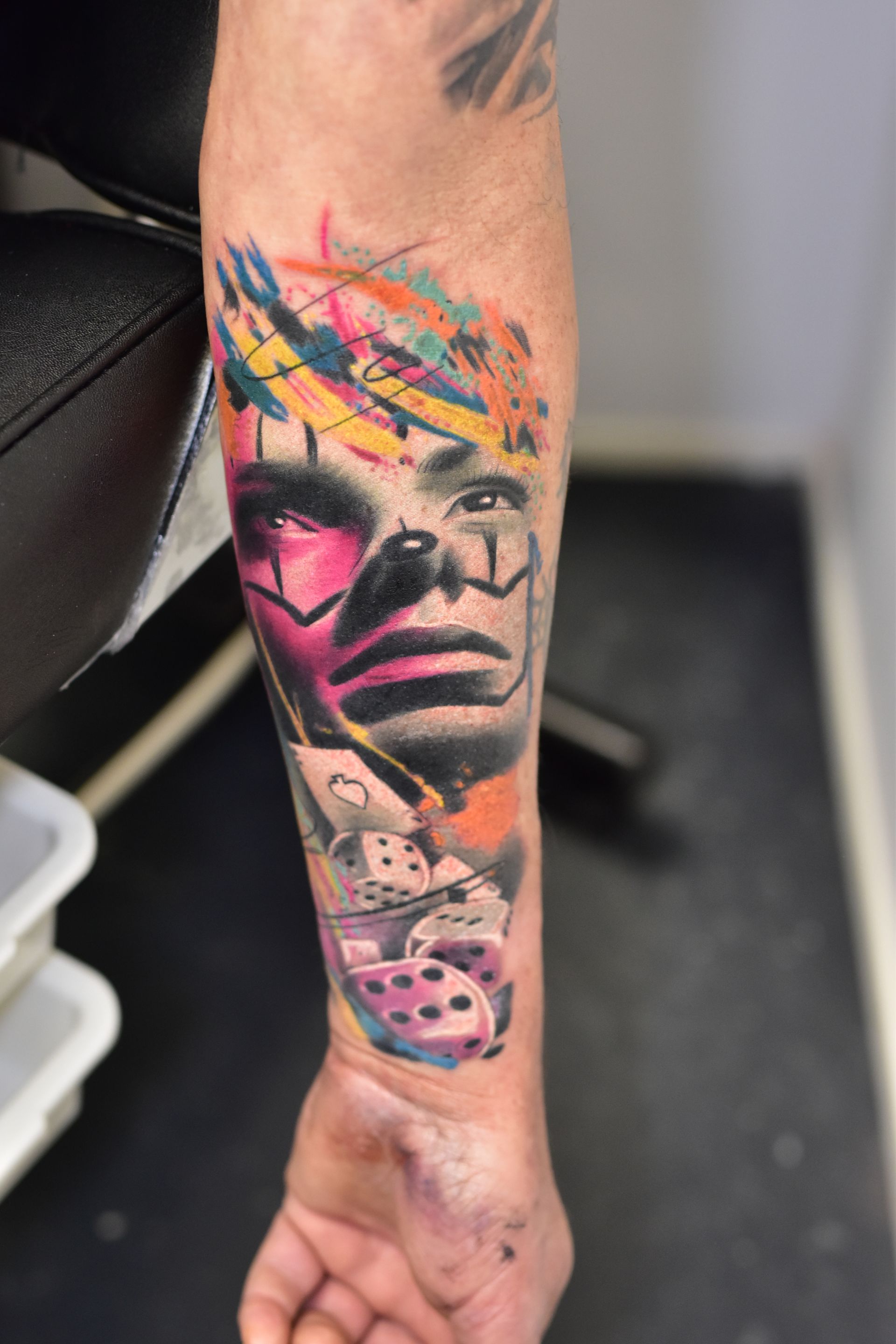 Een tatoeage op de onderarm met een gestileerd portret in levendige penseelstreken, aangevuld met motieven van dobbelstenen en speelkaarten.