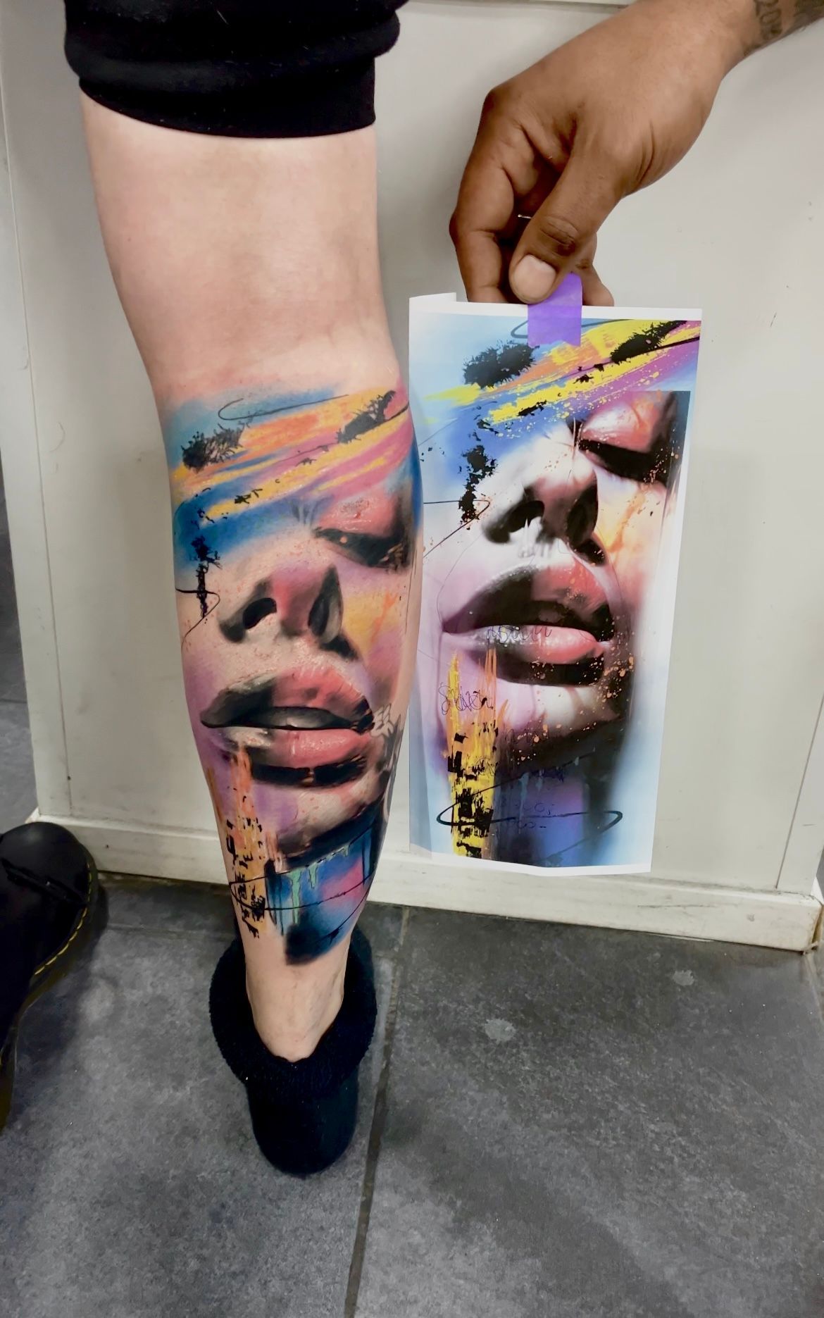 Een tatoeage van een gezicht in een kleurrijke, abstracte aquarel-stijl op het been van een persoon, afgebeeld naast het referentieontwerp.