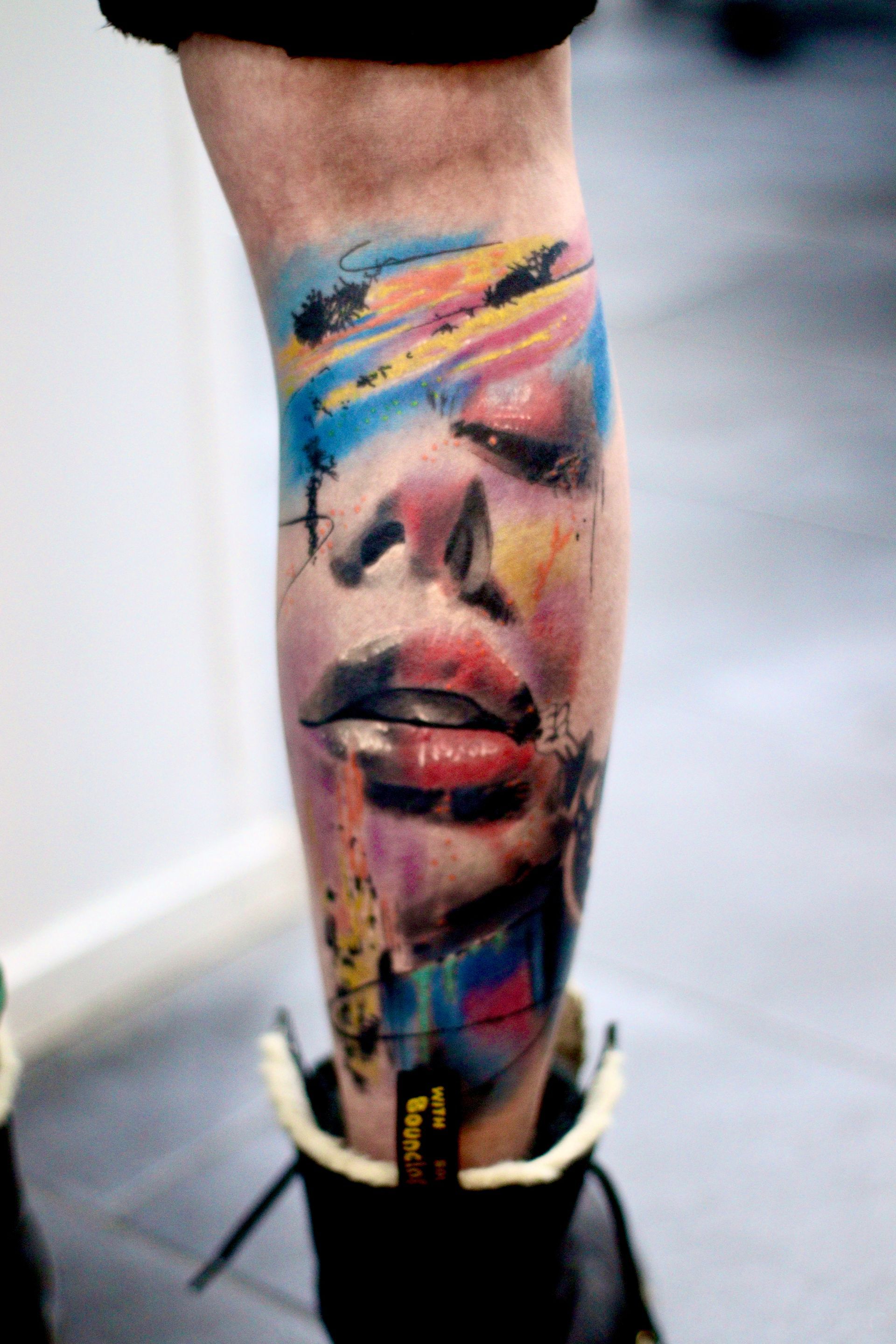Abstracte portrettattoo in aquarel-stijl op een onderbeen met levendige blauwe, rode en gele penseelstreken.