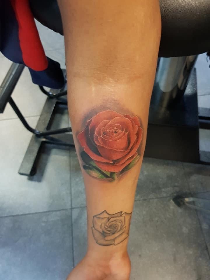 Een onderarm met een levendige, rode, realistische roos-tatoeage boven een kleinere, zwart getinte roos-tatoeage met contouren.