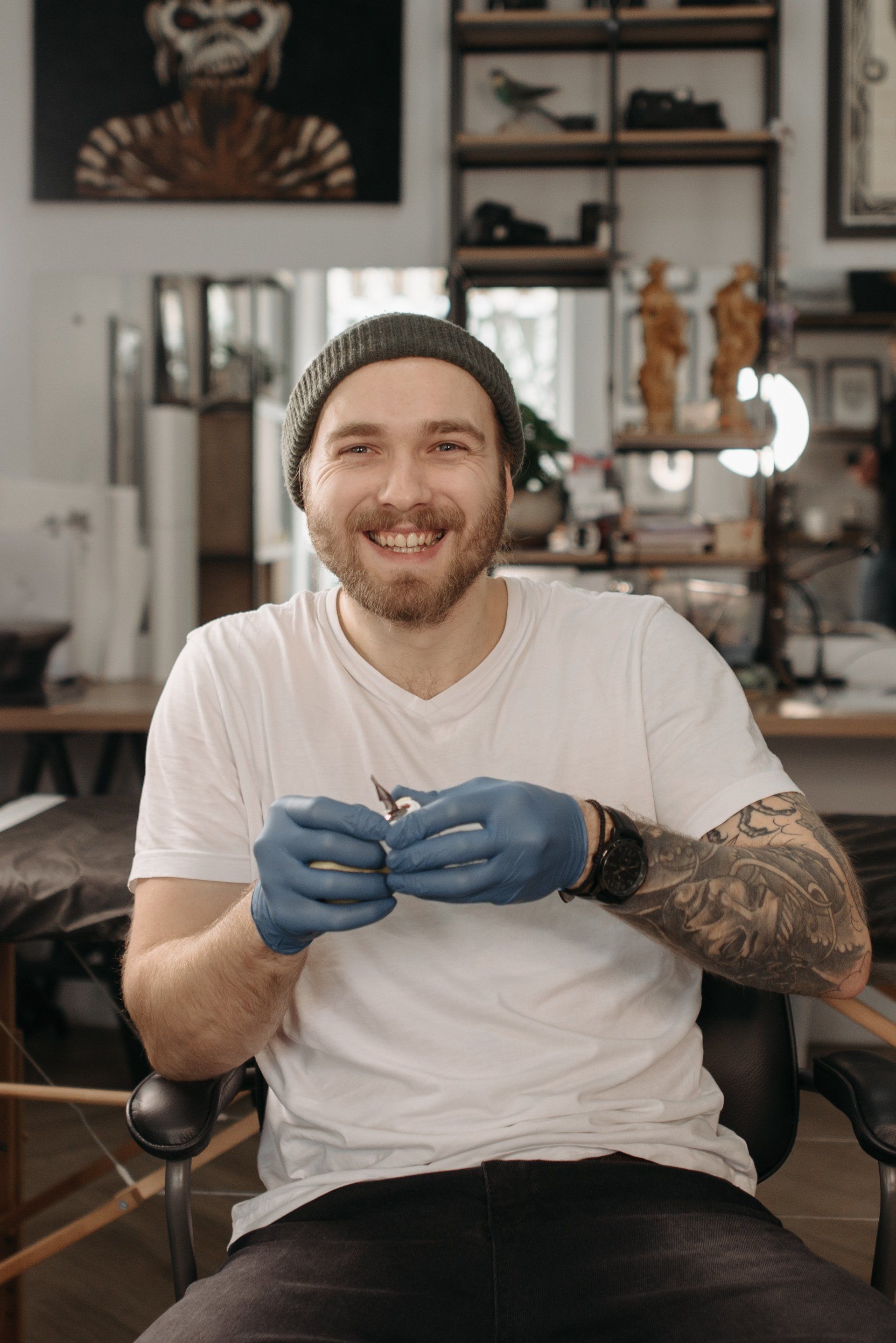 Een lachend persoon met een muts en een wit T-shirt, getatoeëerde armen en blauwe medische handschoenen, aan het werk in een studio.