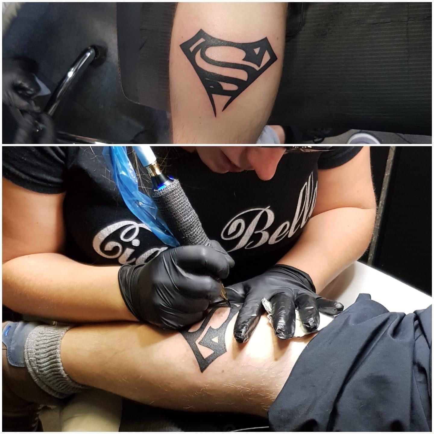 Een tatoeëerder met zwarte handschoenen werkt aan een zwarte Superman-tatoeage op de kuit van een persoon, zoals te zien is in twee aanzichten.