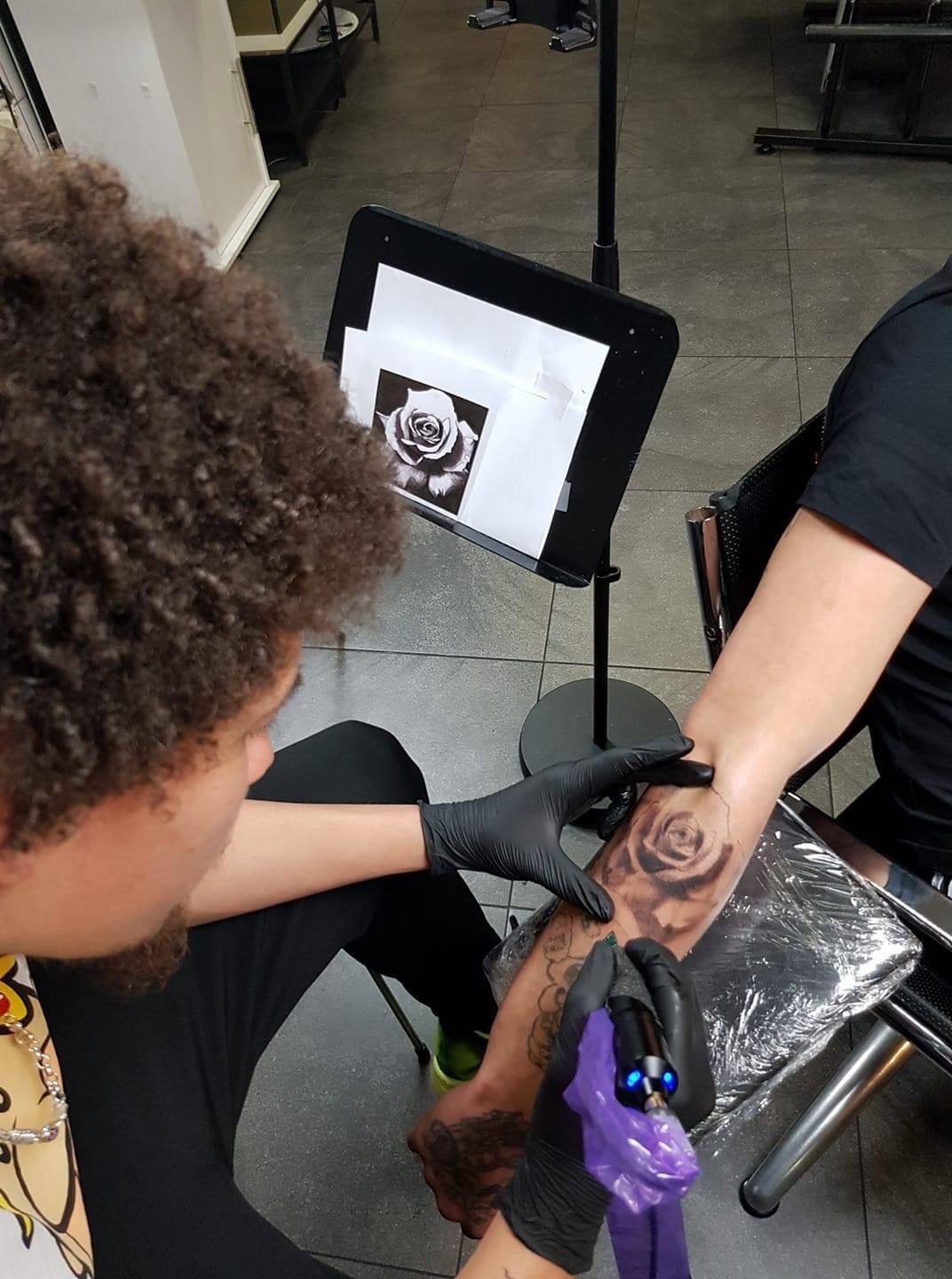 In een studio gebruikt iemand met zwarte handschoenen een tatoeëermachine om een ​​roos op de arm van een andere persoon te tatoeëren.