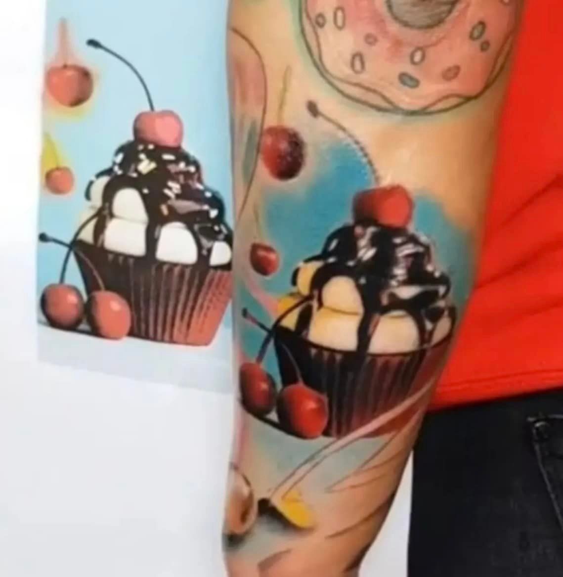 Een tatoeage op de onderarm van een cupcake met chocolade en kersen, afgebeeld naast een voorbeeldtekening op een blauwe achtergrond.