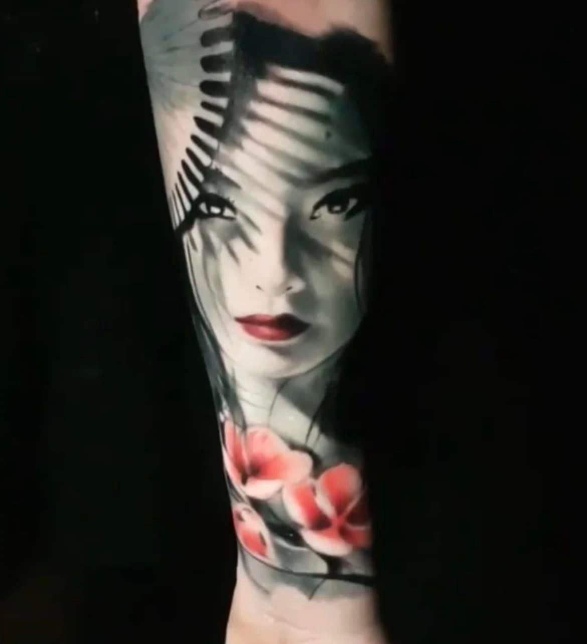 Een tatoeage van een gezicht dat in de schaduw van een waaier ligt, met kleine roze bloemen onderaan op een zwarte achtergrond.