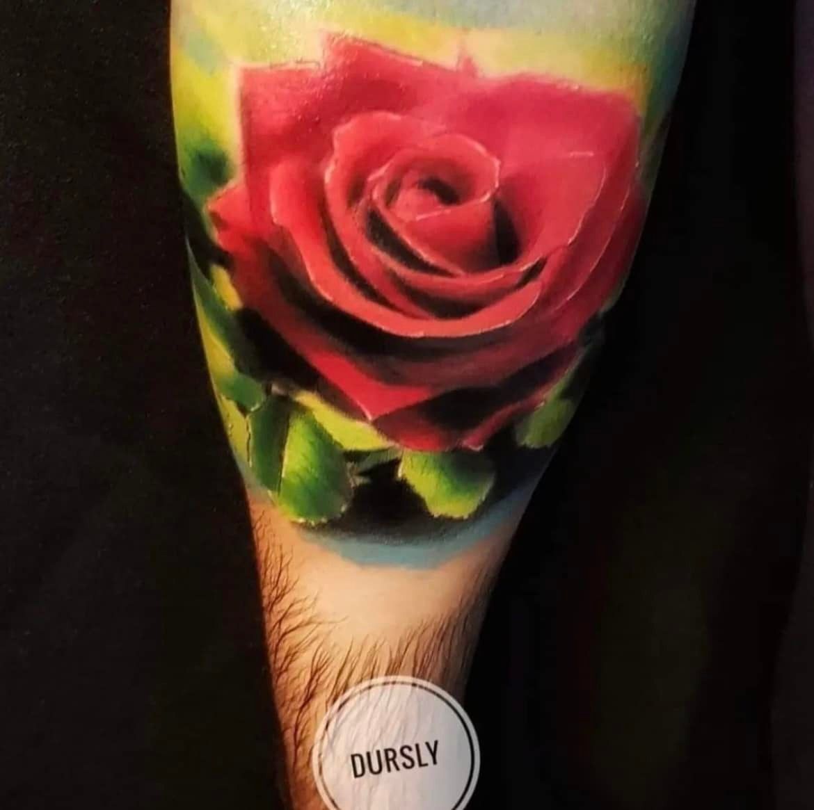 Een levendige rode roos-tatoeage op de huid, met zachtgroene bladeren en een wazige geelblauwe achtergrond.