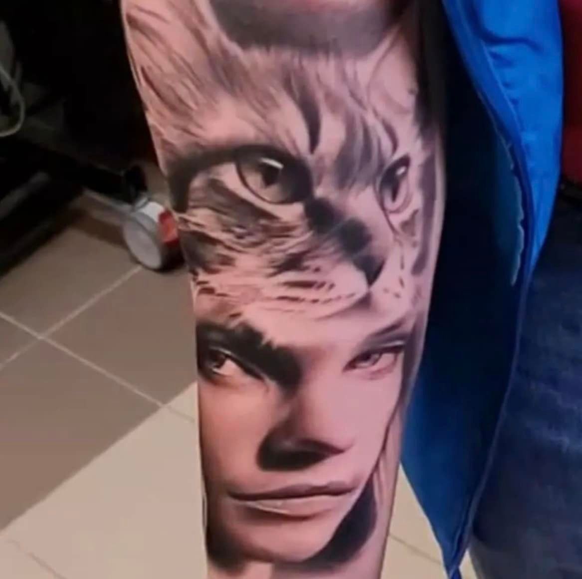 Een zwart-grijze tatoeage op een arm met een realistisch kattengezicht dat naadloos overgaat in het gezicht van een persoon.
