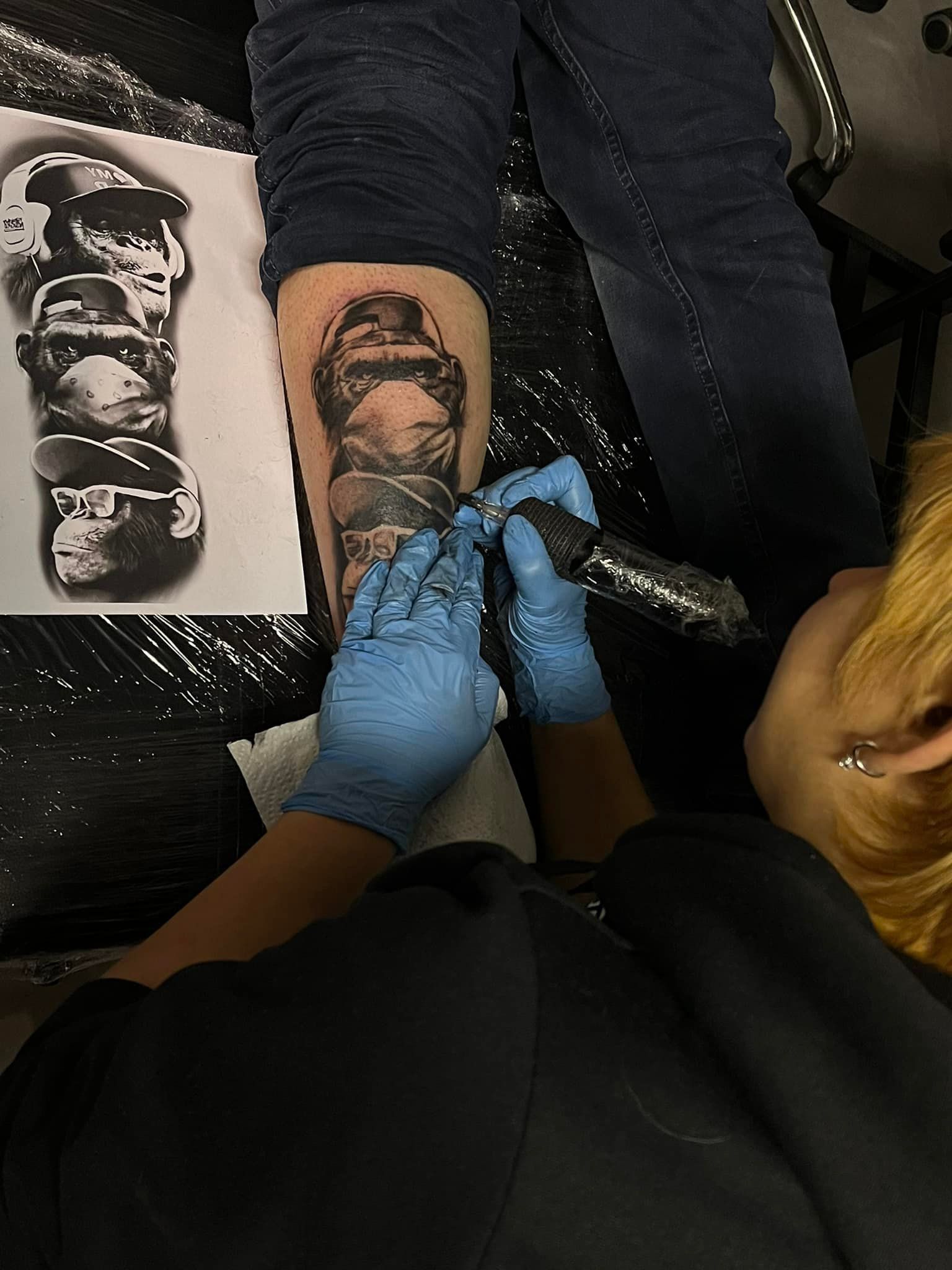 Een tatoeëerder met blauwe handschoenen gebruikt een roterende machine om een ​​gorilla op het onderbeen van een persoon te tatoeëren.