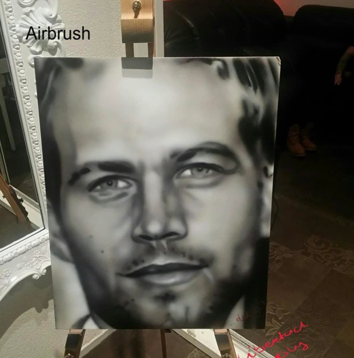 Een zwart-wit portret, geschilderd met airbrush, van een man met lichte ogen op een doek dat op een schildersezel staat.