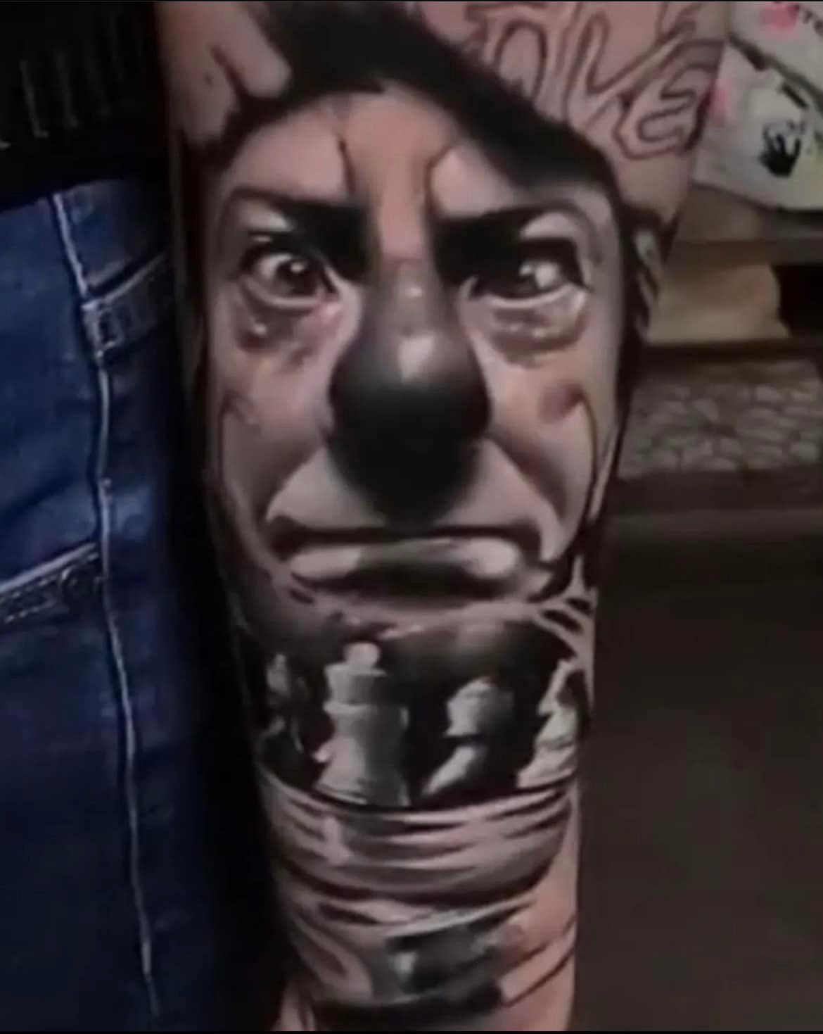 Een gedetailleerde zwart-grijze tatoeage op een onderarm met een clowngezicht en schaakstukken aan de onderkant.