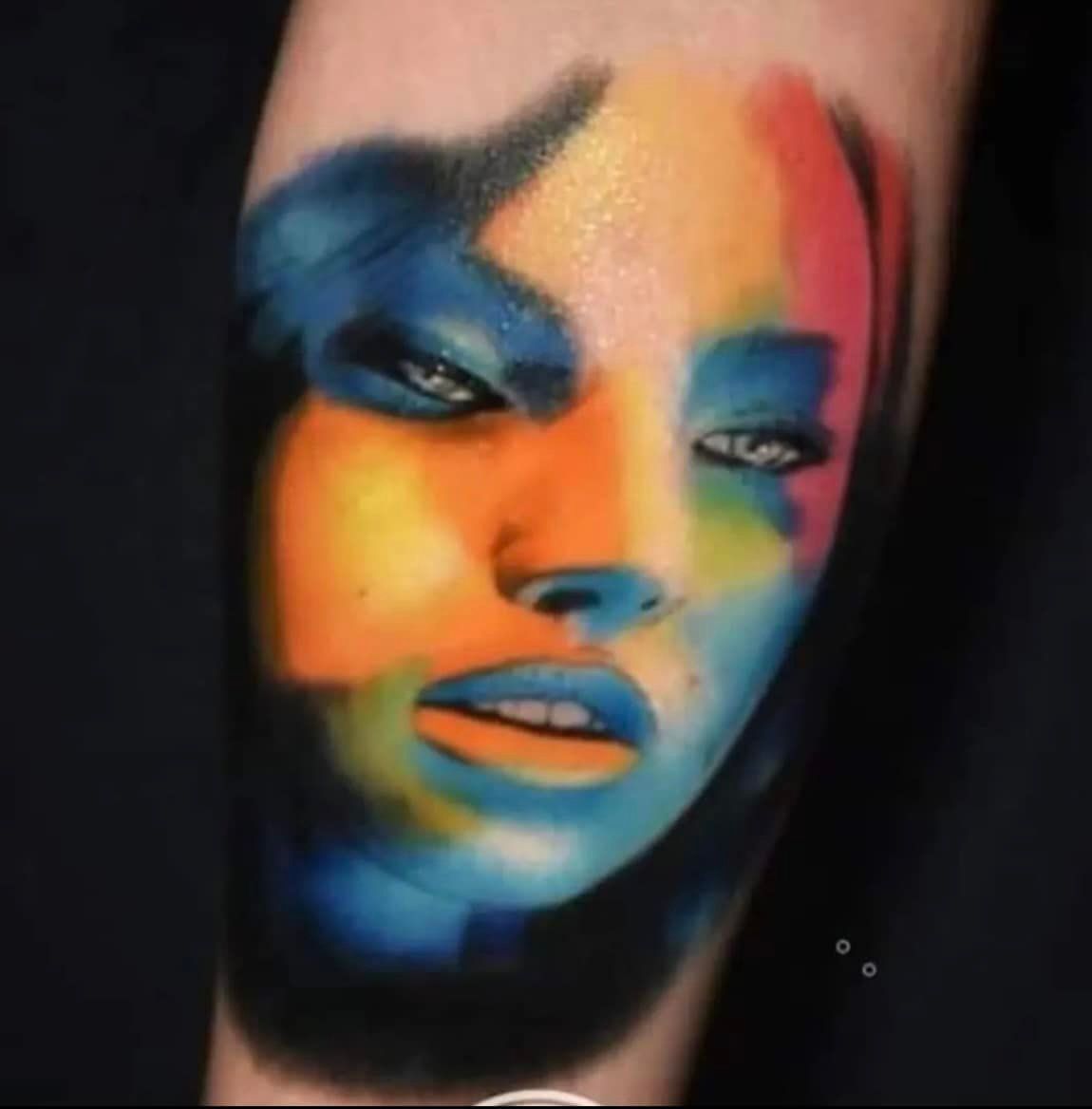 Close-up van een kleurrijke, abstracte portrettattoo op de huid, met een mengeling van blauwe, oranje en rode tinten.