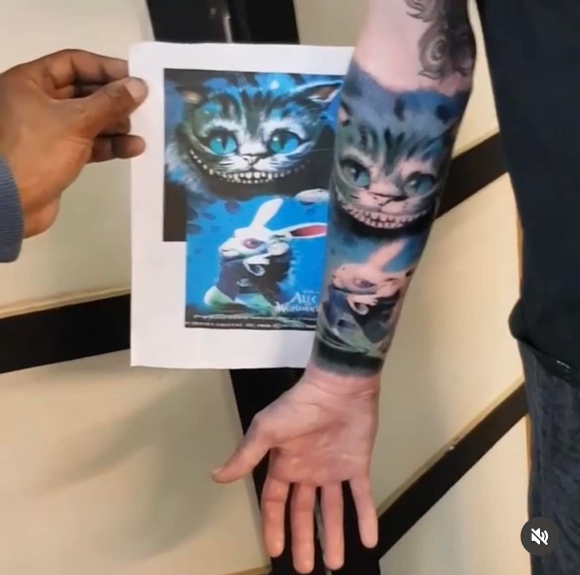 Een hand houdt een afdruk van de Cheshire Cat en het Witte Konijn vast, naast een bijpassende tatoeage op de onderarm van een persoon.
