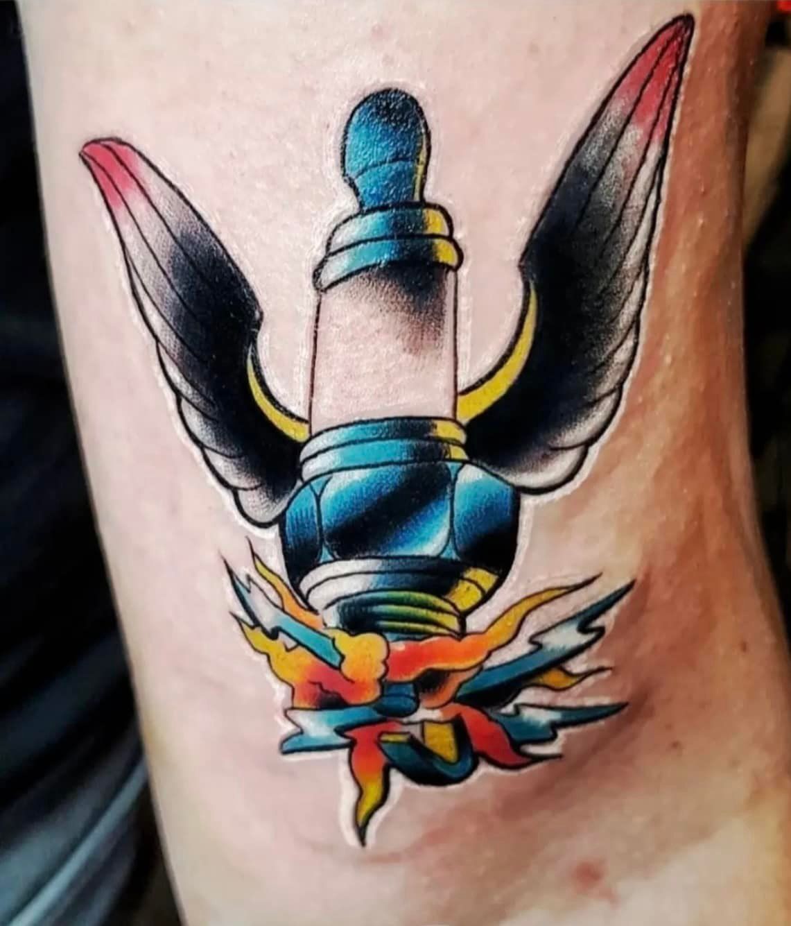 Een neotraditionele tatoeage van een gevleugelde bougie met vlammen, uitgevoerd in zwart, blauw, rood en geel op de huid.