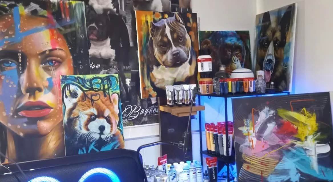 Een verzameling kleurrijke portret- en dierenschilderijen tentoongesteld in een kunstenaarsatelier, met benodigdheden op een metalen plank.