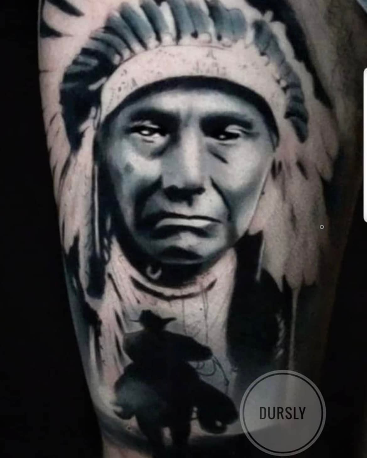 Een zwart-grijze tatoeage van een inheemse persoon met een verentooi, met daaronder een klein silhouet van een ruiter.