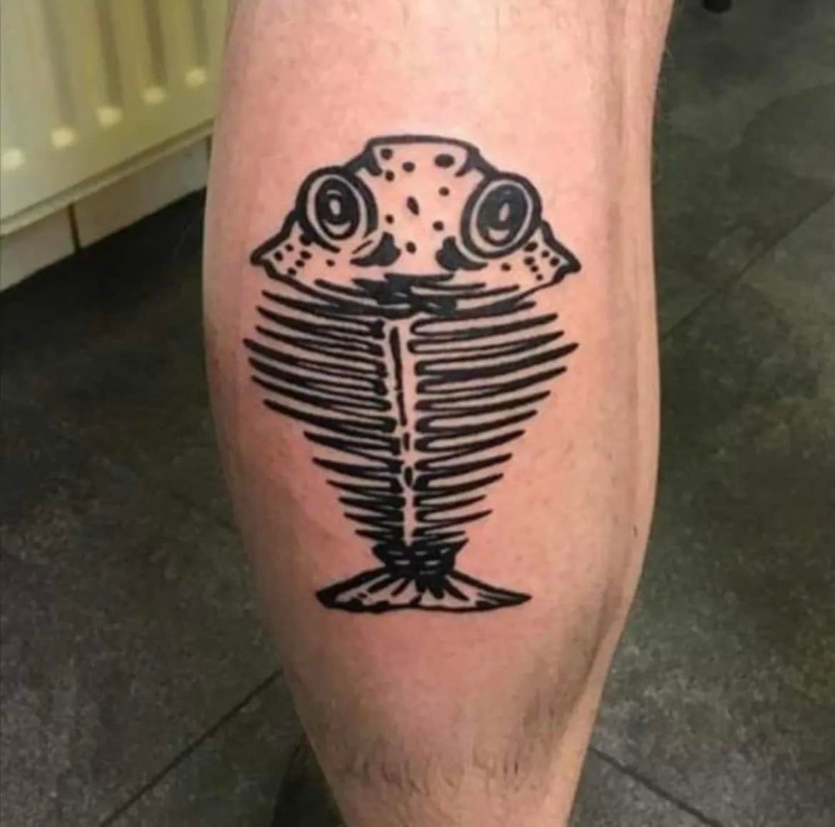 Een opvallende zwarte tatoeage op iemands been, die een cartoonachtig, wijdogig visskelet voorstelt.
