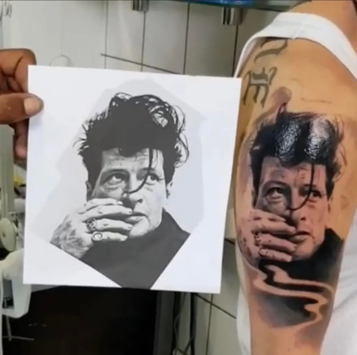 Een hand houdt een referentiefoto van een persoon vast naast een realistische portrettattoo van diezelfde persoon op een arm.
