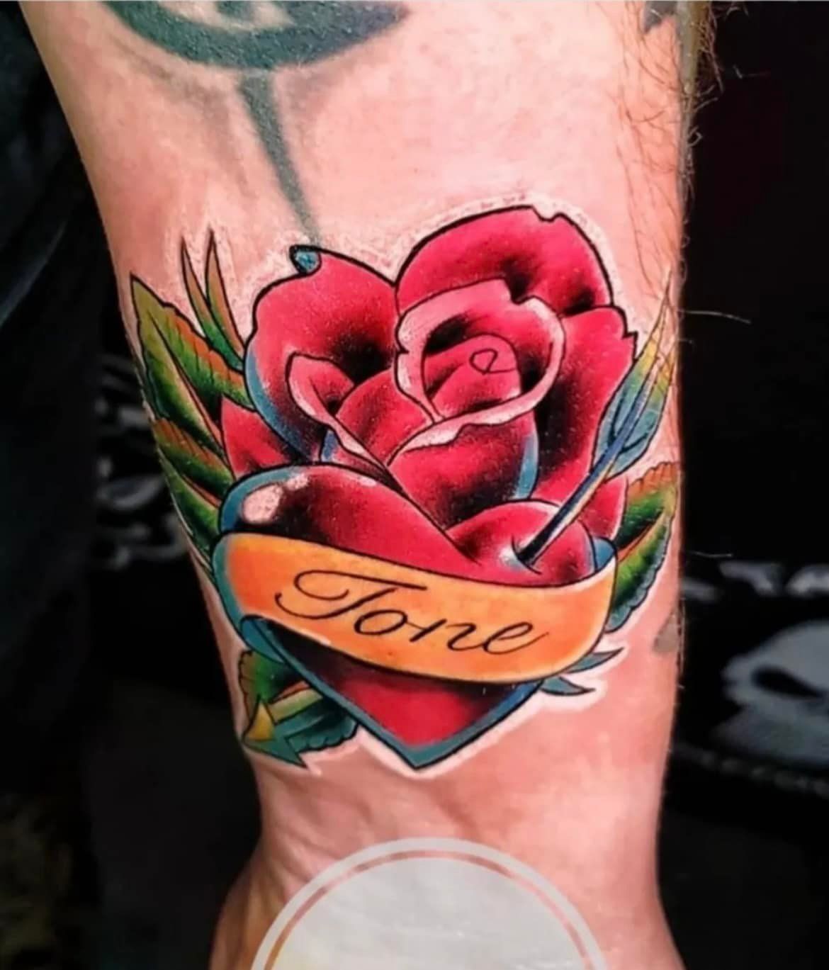 Een kleurrijke tatoeage op een arm met een rode roos, een kleine blauwe pijl en een gouden banner met de naam