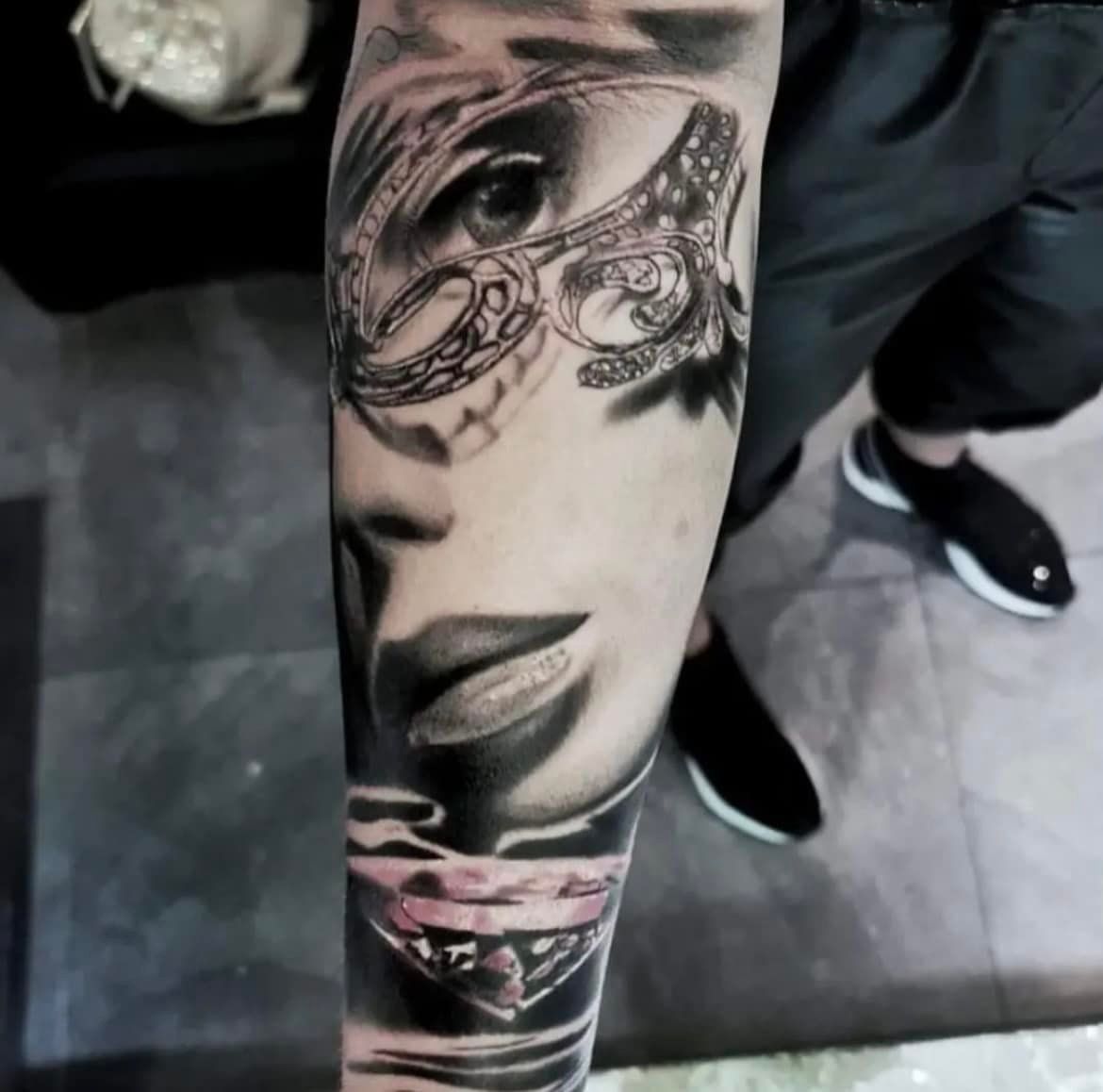 Een zwart-grijze tatoeage op de onderarm van een gezicht met een sierlijk, gedessineerd masker, met een ruitvorm onder de lippen.
