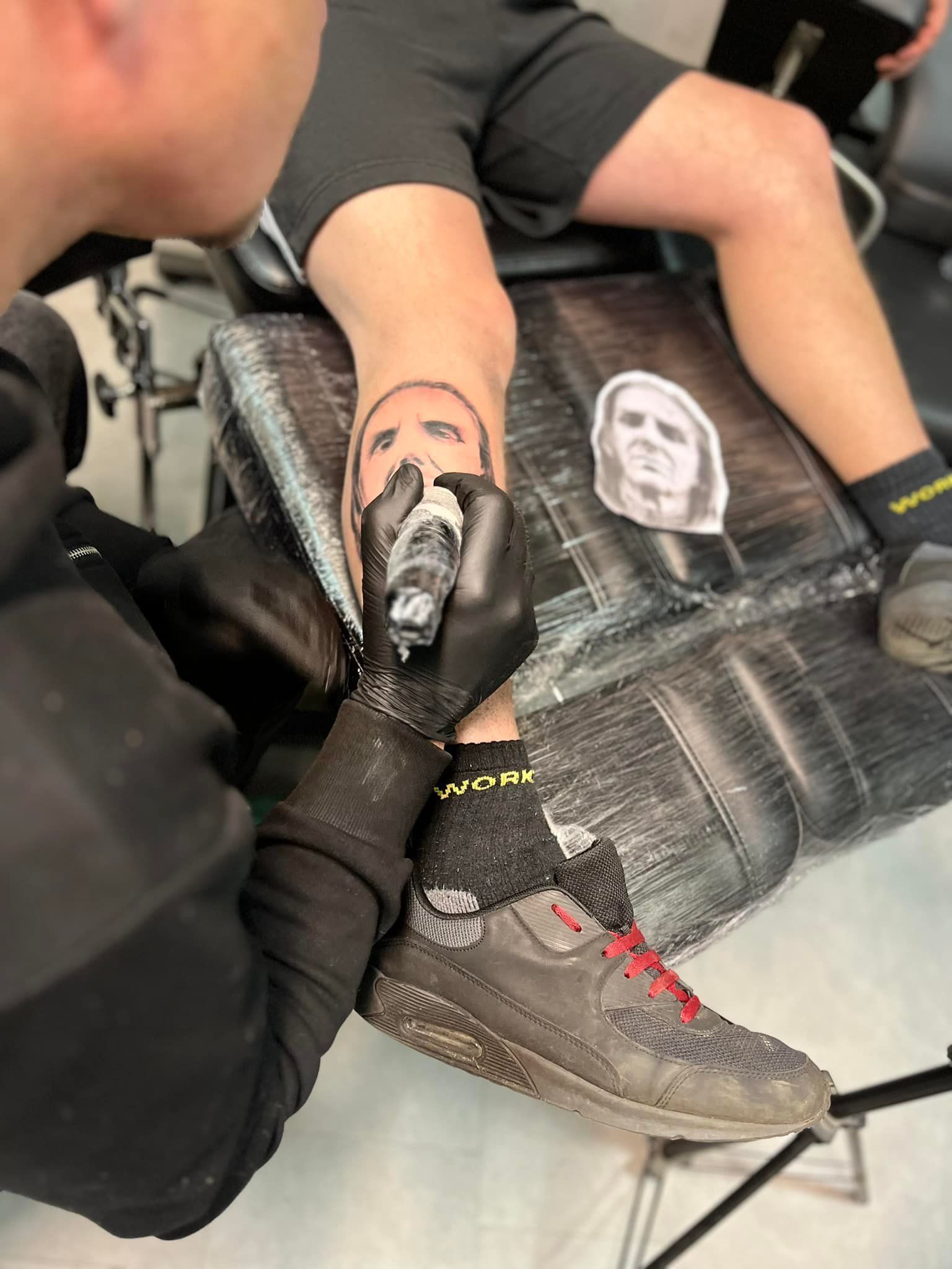 Een tatoeëerder gebruikt een machine om een ​​portret op het onderbeen van een persoon te tatoeëren, aan de hand van een referentiefoto die ernaast is geplaatst.