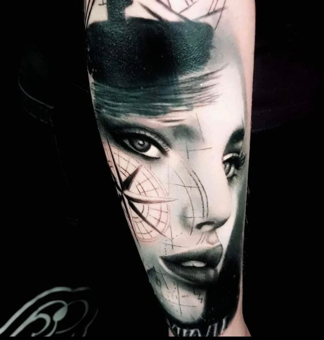 Een zwart-grijze tatoeage op de huid, die een gezicht afbeeldt met een kompasmotief eroverheen, waarbij schetsachtige lijnen en schaduwen in elkaar overlopen.