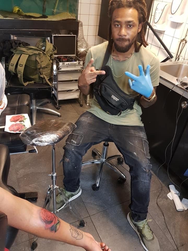 Een persoon met dreadlocks zit in een tattoostudio, gekleed in een groen T-shirt, een zwarte broek en een blauwe handschoen, terwijl hij poseert.