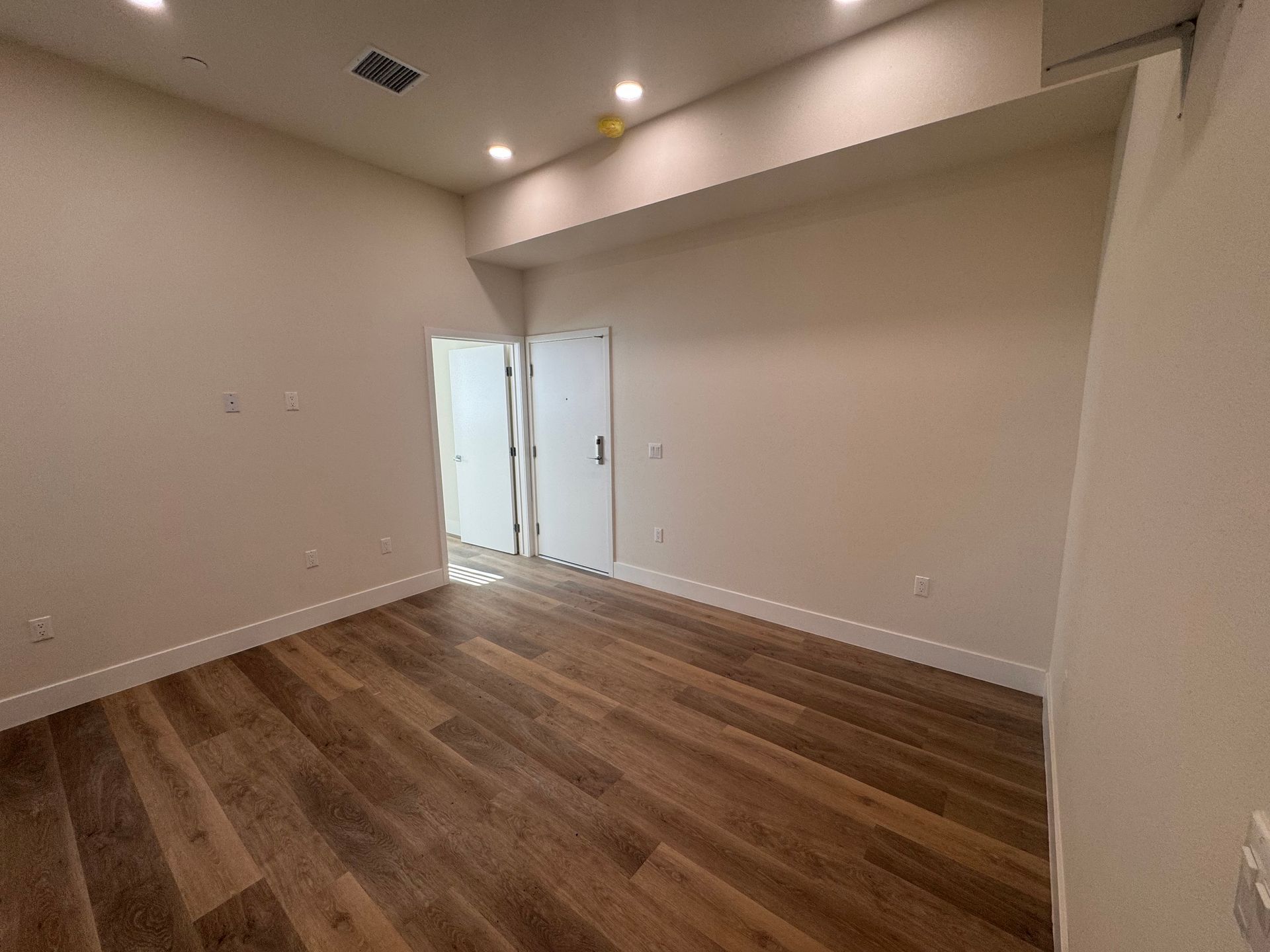 An empty, modern room with light-colored walls, wood-look flooring, recessed lighting, and two white doors in the back.