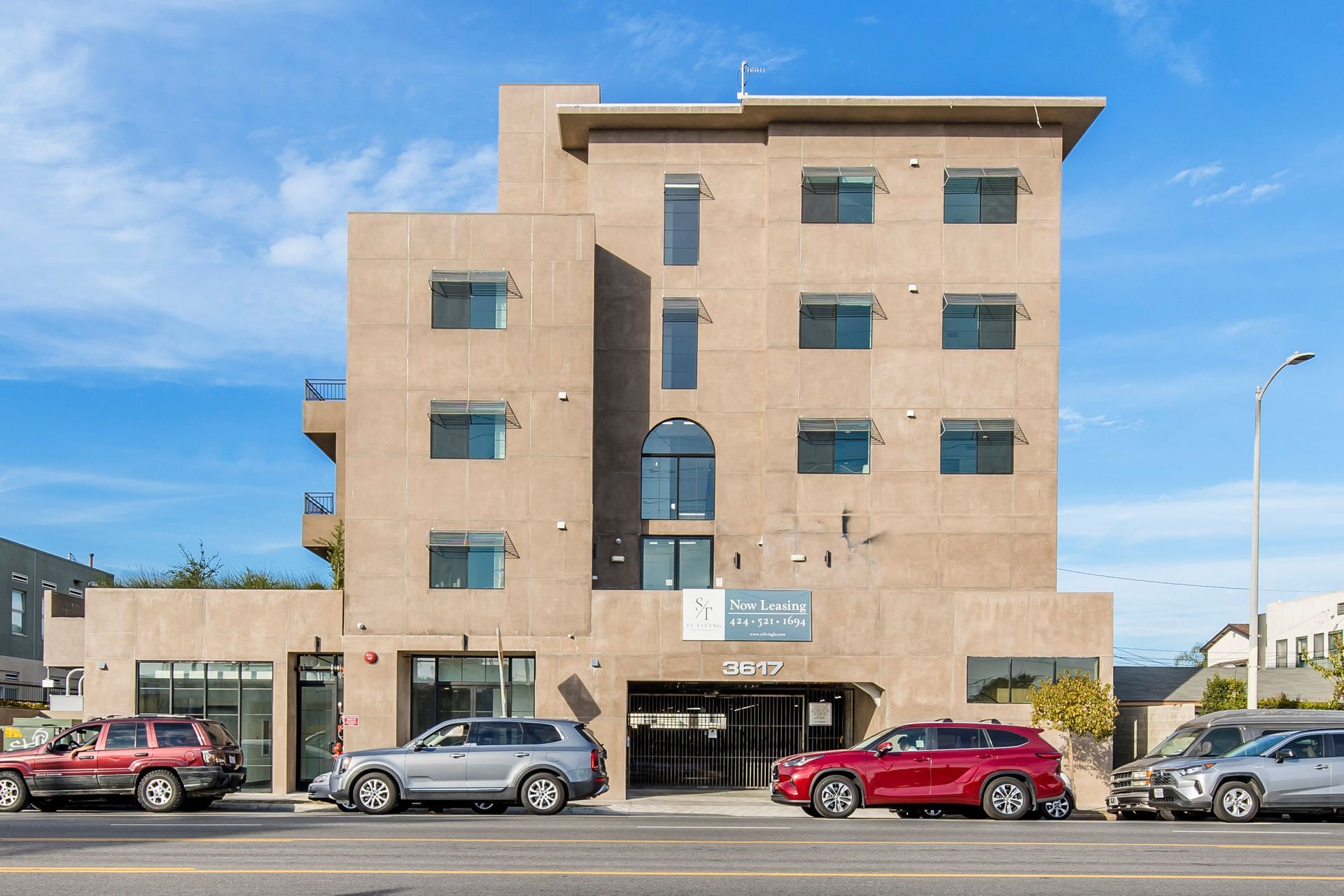 A multi-story tan stucco apartment building with a ground-level parking garage and several cars parked in front.