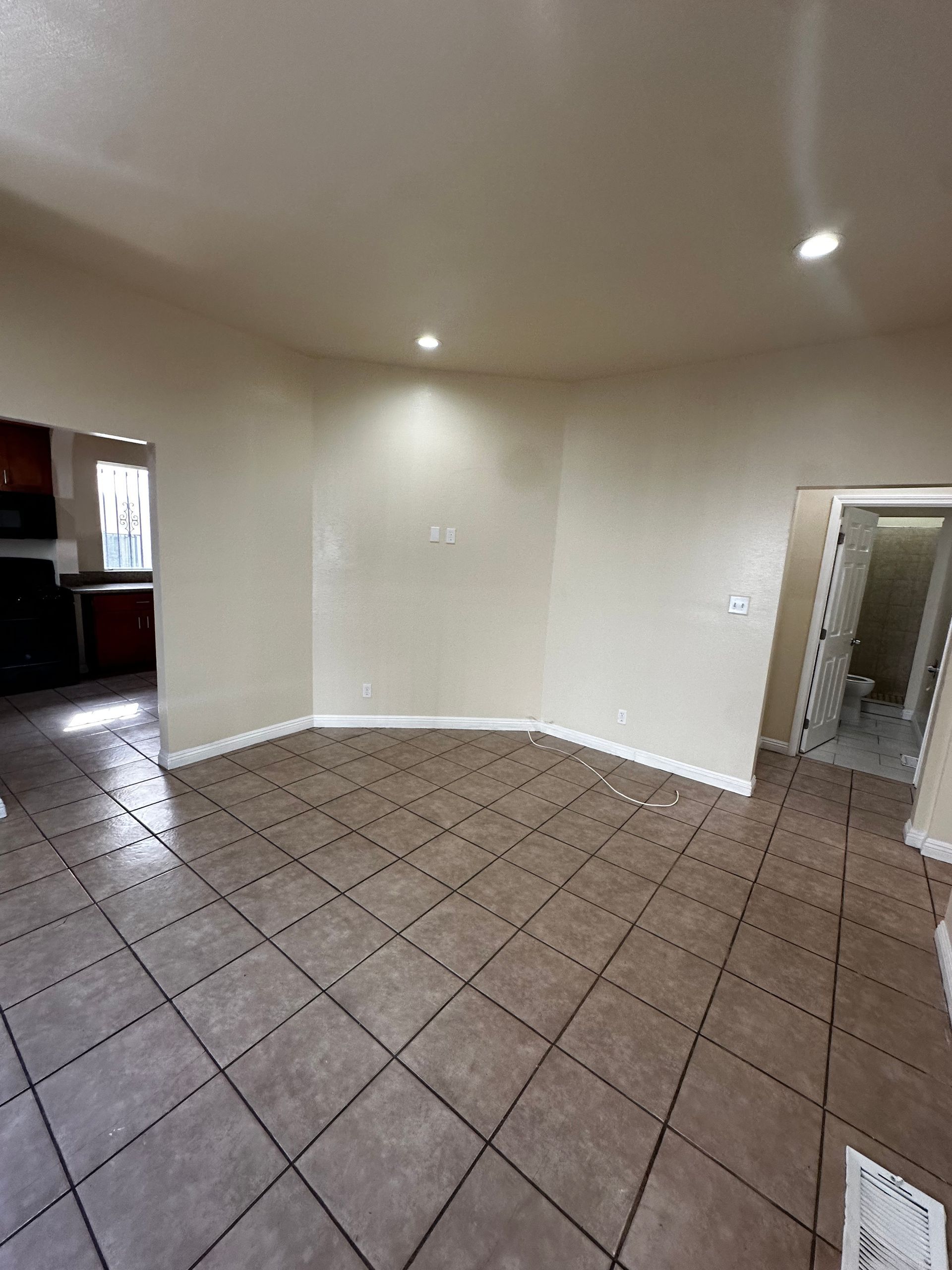 An empty room with tan walls, brown square-tiled flooring, recessed ceiling lights, and two doorways.