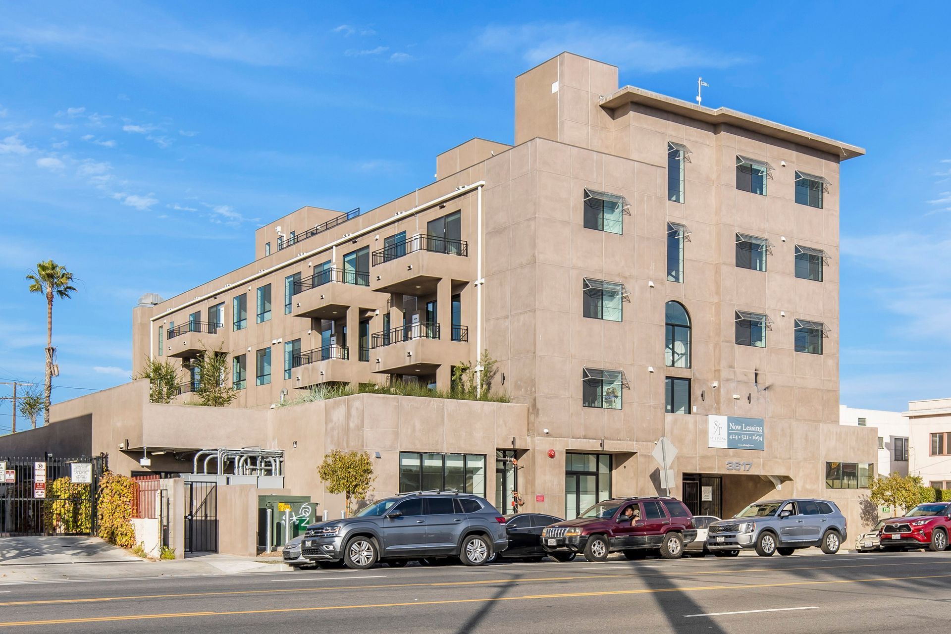 A tan, multi-story apartment building with balconies and large windows, fronted by a parking row of cars on a sunny day.