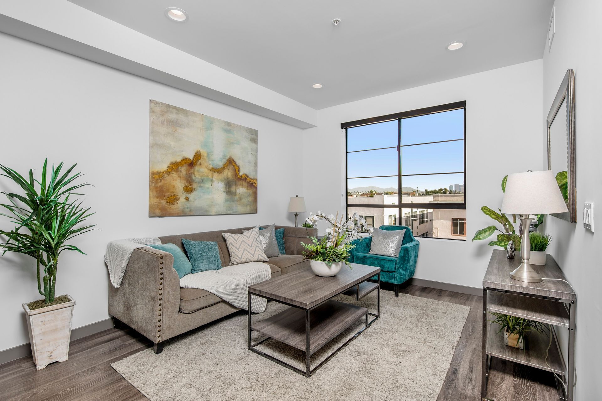 A modern, minimalist living room with a gray sectional sofa, teal armchair, wooden coffee table, and large window.