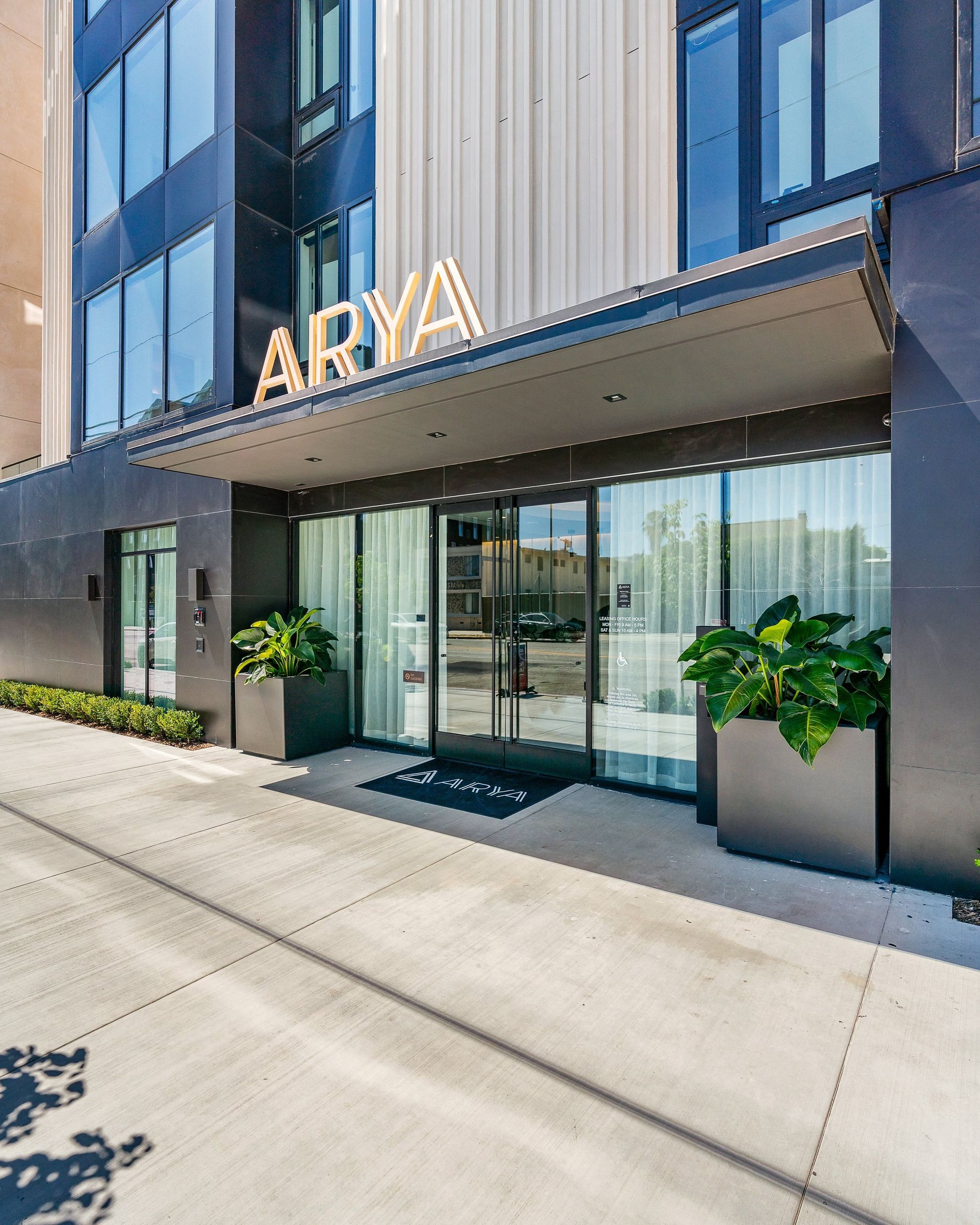 The entrance to the Arya building, featuring a dark exterior, a modern sign, glass doors, and two large potted plants.