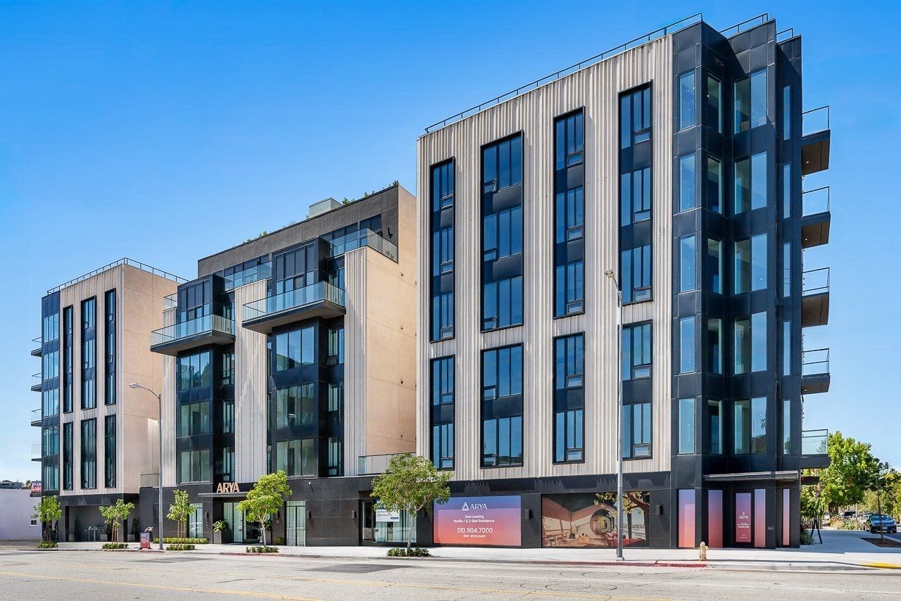 A modern apartment complex with tan and dark grey panels, large windows, and street-level storefronts under a blue sky.