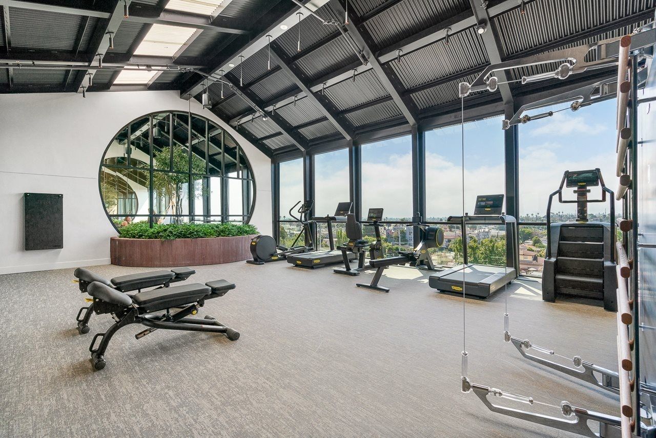 A bright, modern gym with workout benches, treadmills, and a circular window overlooking a city view.