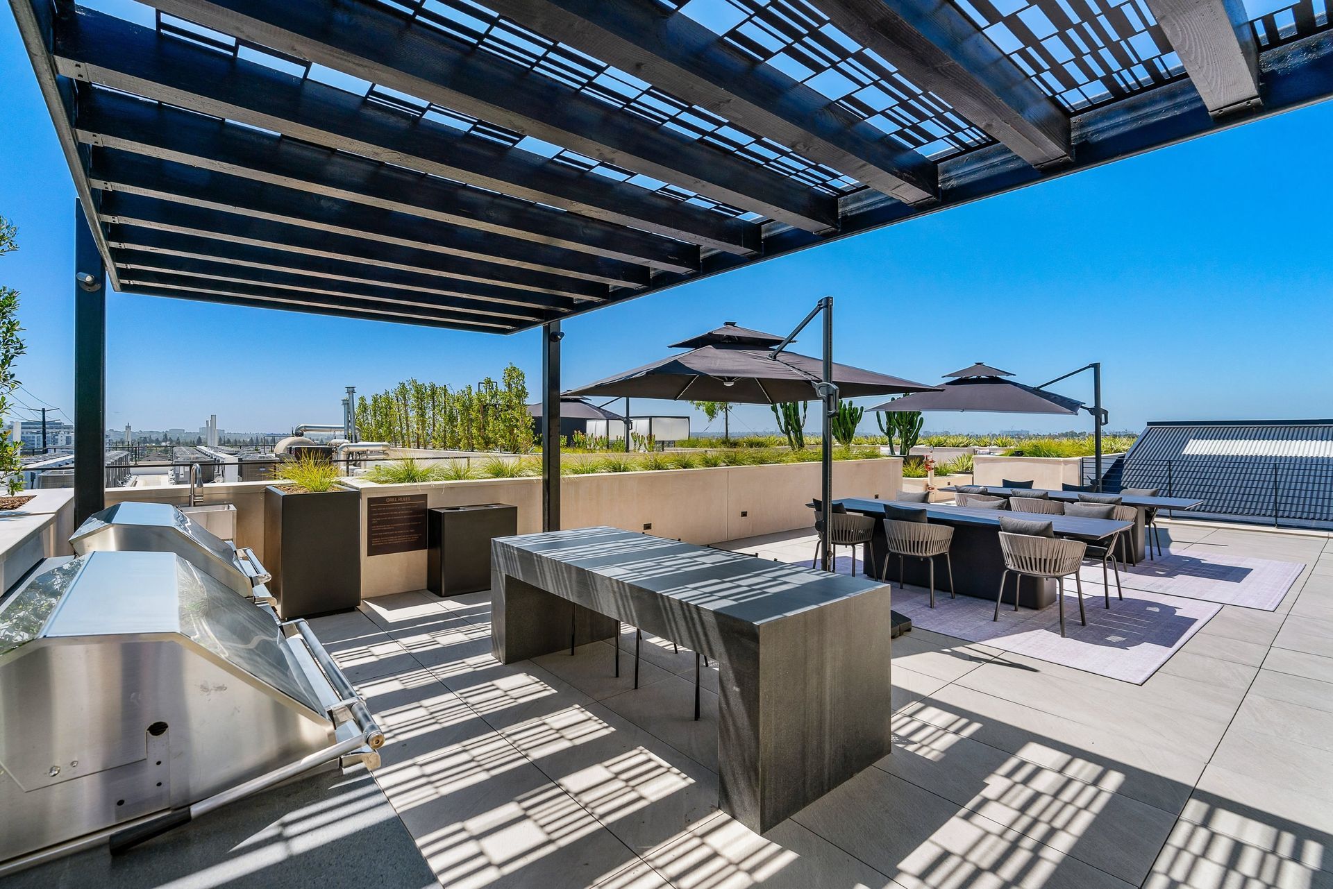 A rooftop patio featuring a stainless steel grill, a stone island, and dining tables under a shaded modern pergola.