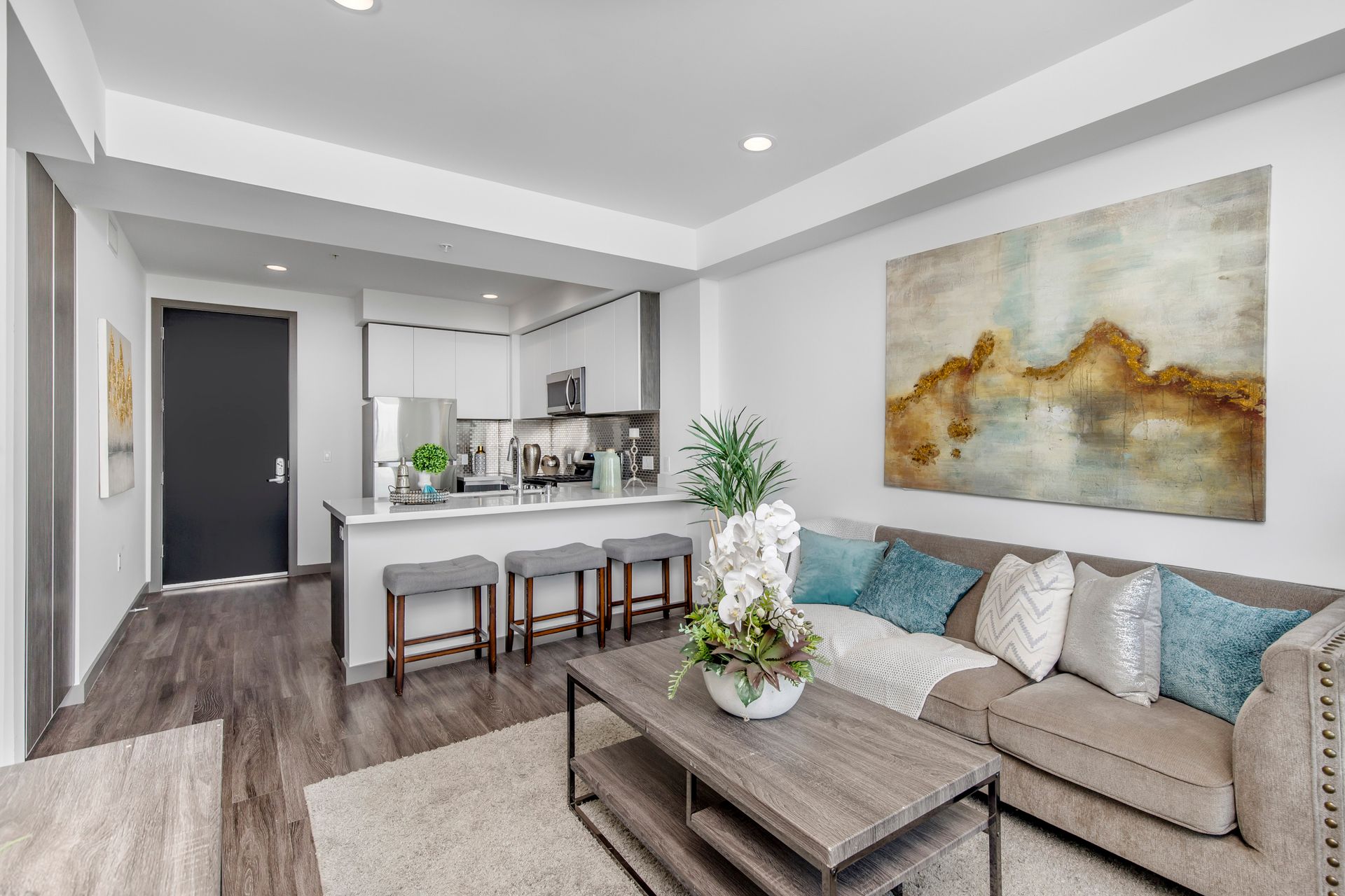 A brightly lit apartment living room with a neutral sofa, coffee table, and an open kitchen with a breakfast bar.