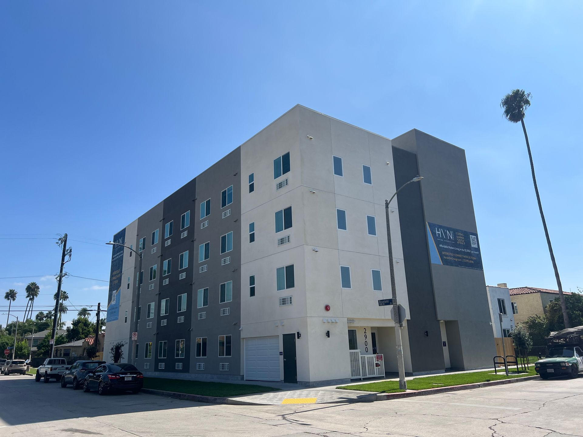 A four-story modern apartment building with white and dark gray panels, parked cars, and a palm tree under a clear sky.